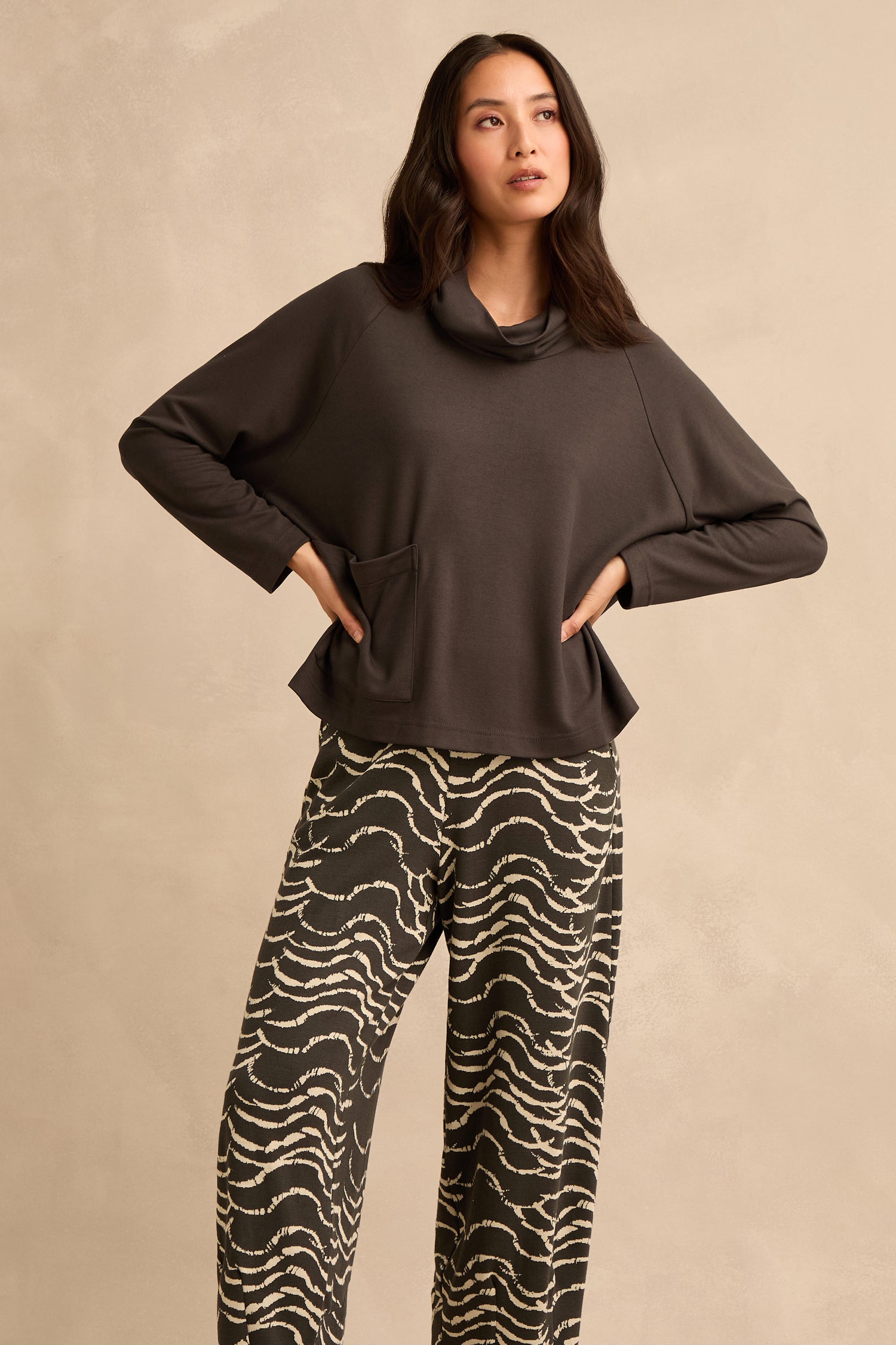 A woman stands against a beige background, wearing the Luxe Viscose Jersey Cowl Neck Top—a loose dark gray style with a front pocket—paired with black-and-white wavy patterned wide-leg pants. She poses with hands on hips, looking slightly to the side.