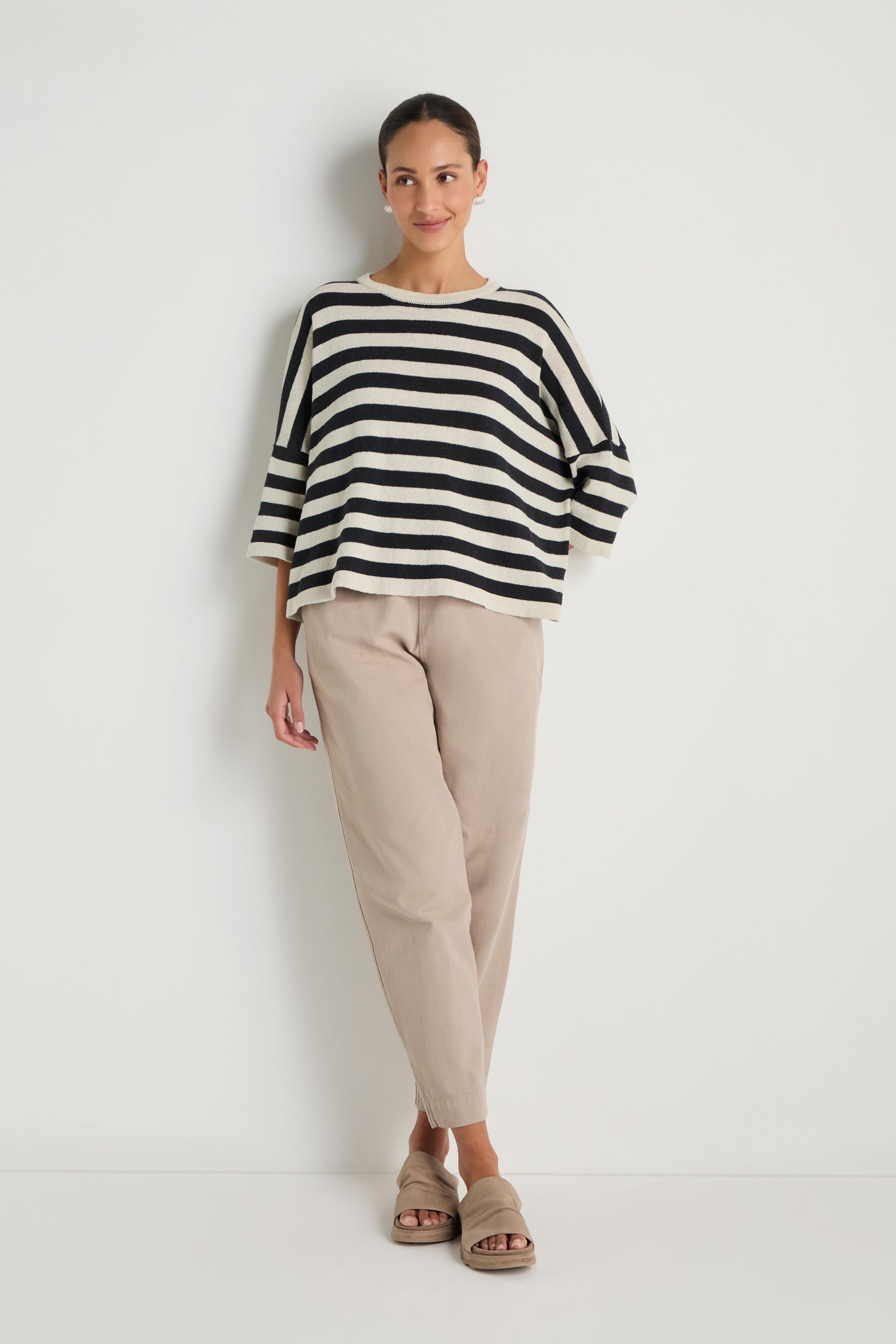 A woman stands against a white wall wearing the Striped Cotton Knit sweater, beige pants, and sandals. Her hair is tied back, and her hands rest relaxed at her sides.