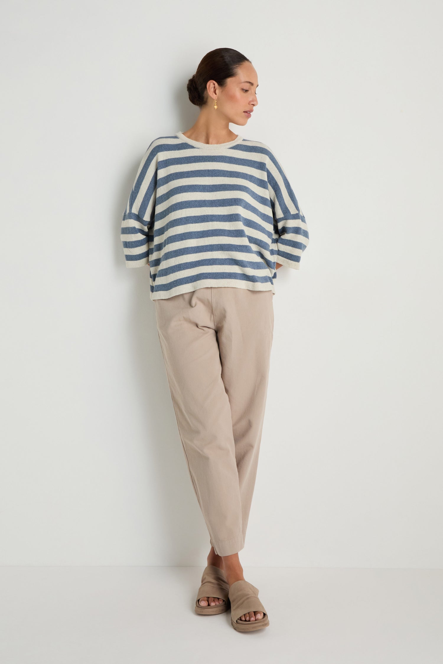 Woman wearing the Striped Cotton Knit—a blue and white sweater—paired with beige pants and sandals, standing against a white wall with hands in pockets. A breathable layer ideal for any casual look.