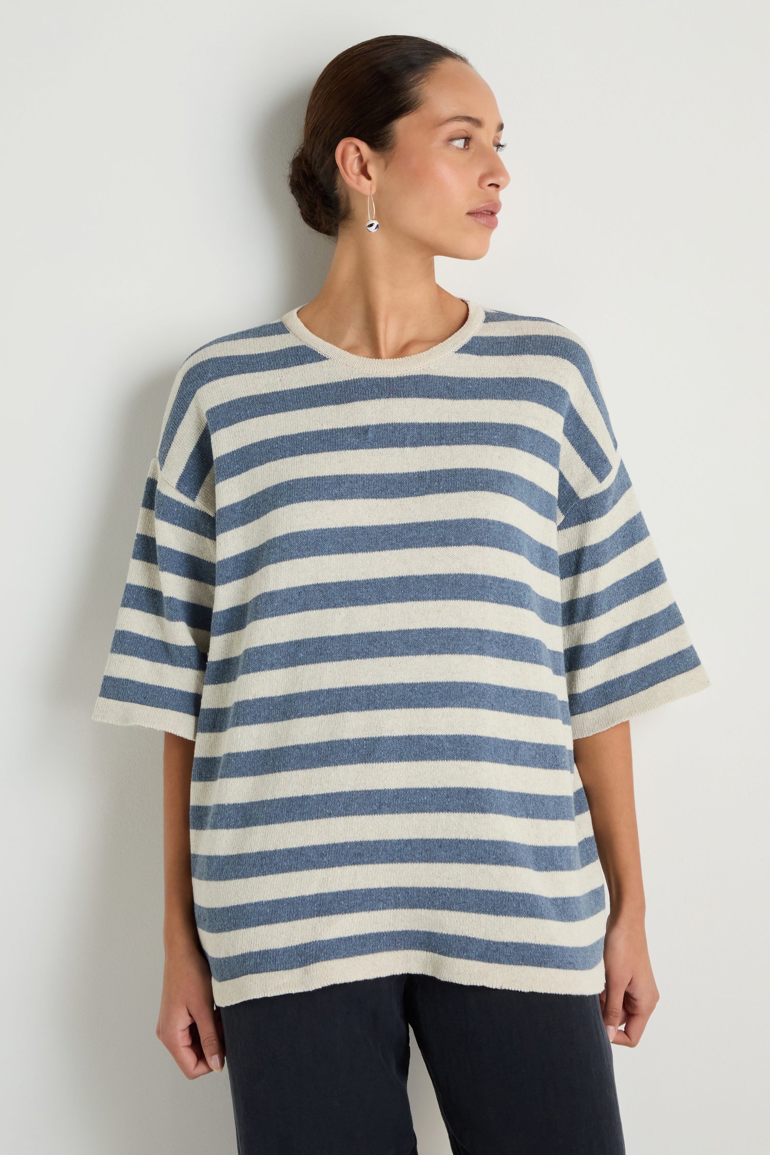 A person stands against a white wall wearing the Striped Cotton Tunic—loose, short-sleeved, blue and white striped cotton—paired with black pants, looking to the side.