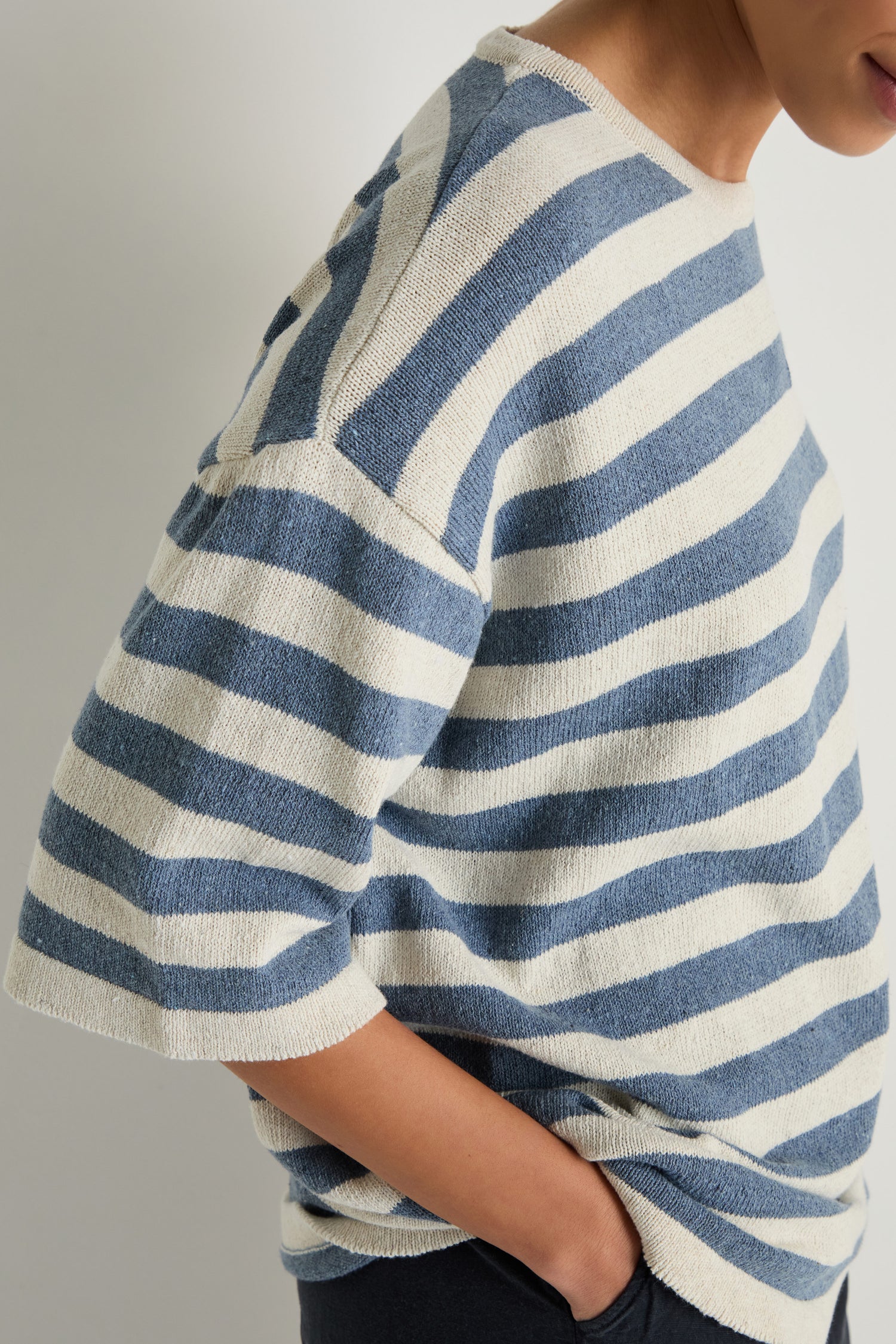 A person is shown from the side with one hand in their pocket, wearing the Striped Cotton Tunic—a loose, short-sleeved top featuring cream and blue stripes.