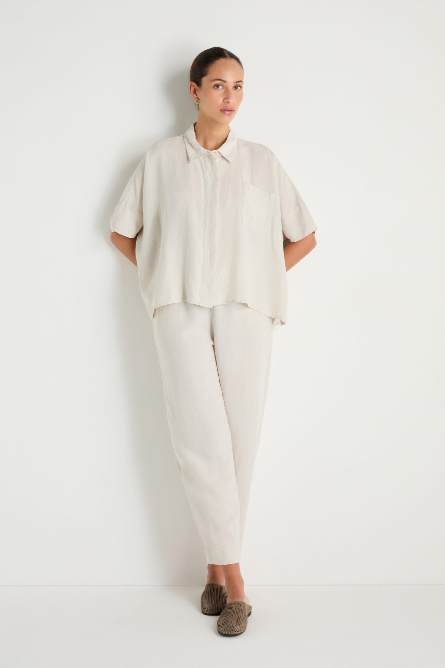 A person stands against a white wall wearing the Lyocell Linen Boxy Shirt in light tones, matching pants, and brown slip-on shoes for an effortlessly casual look.