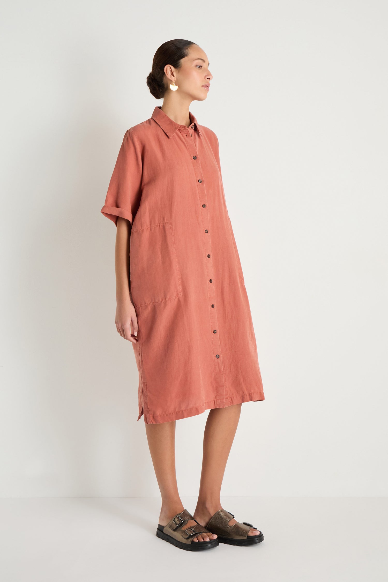 Person stands against a white wall, wearing the Lyocell Linen Shirt Dress—a loose, button-up, short-sleeve rust dress—paired with black sandals and circular earrings.