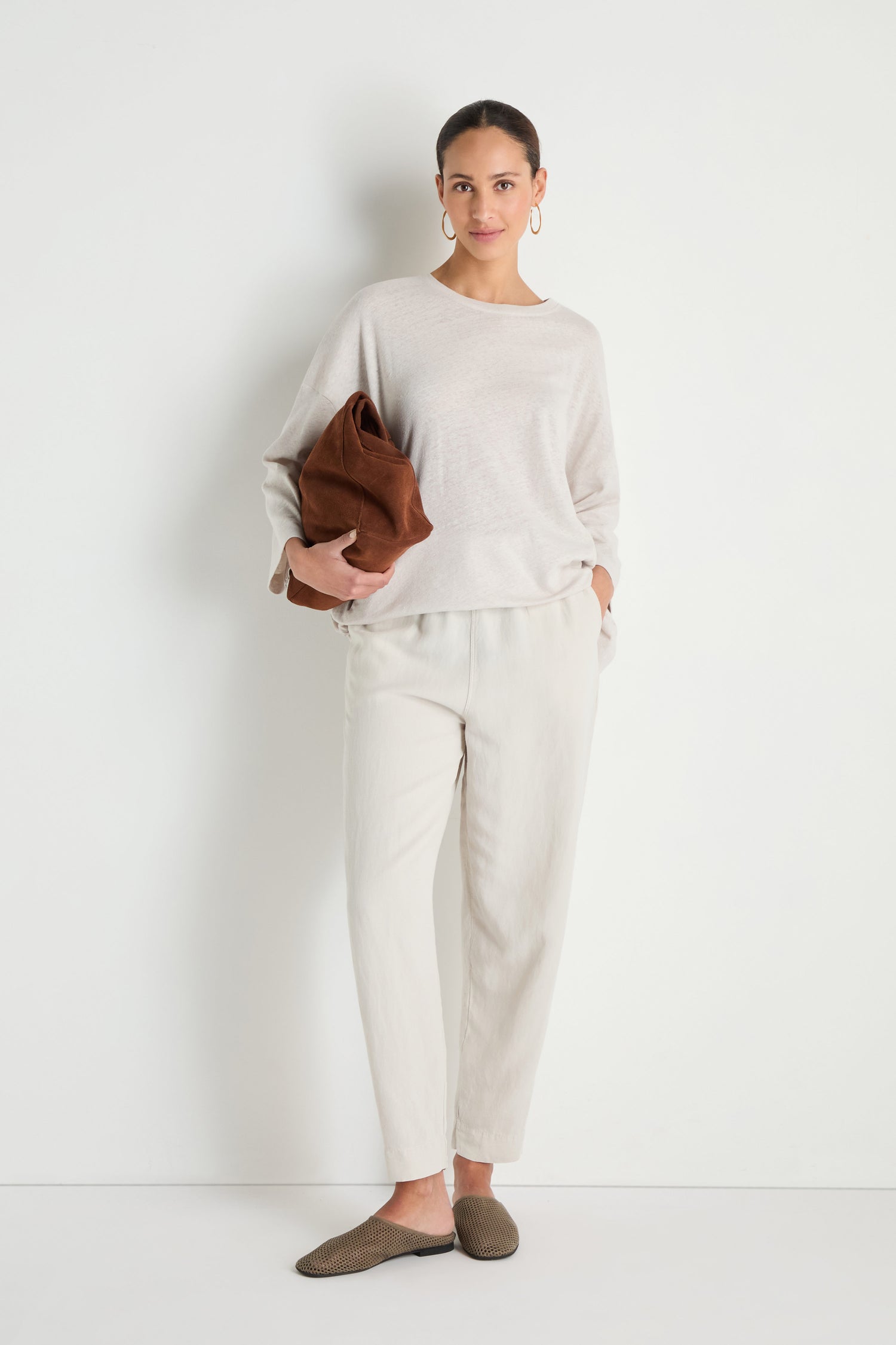 A woman stands against a white wall, wearing a light beige sweater, Cream Lyocell Linen Slim Trousers with an elasticated waist, brown slip-on shoes, hoop earrings, and holding a folded brown bag.