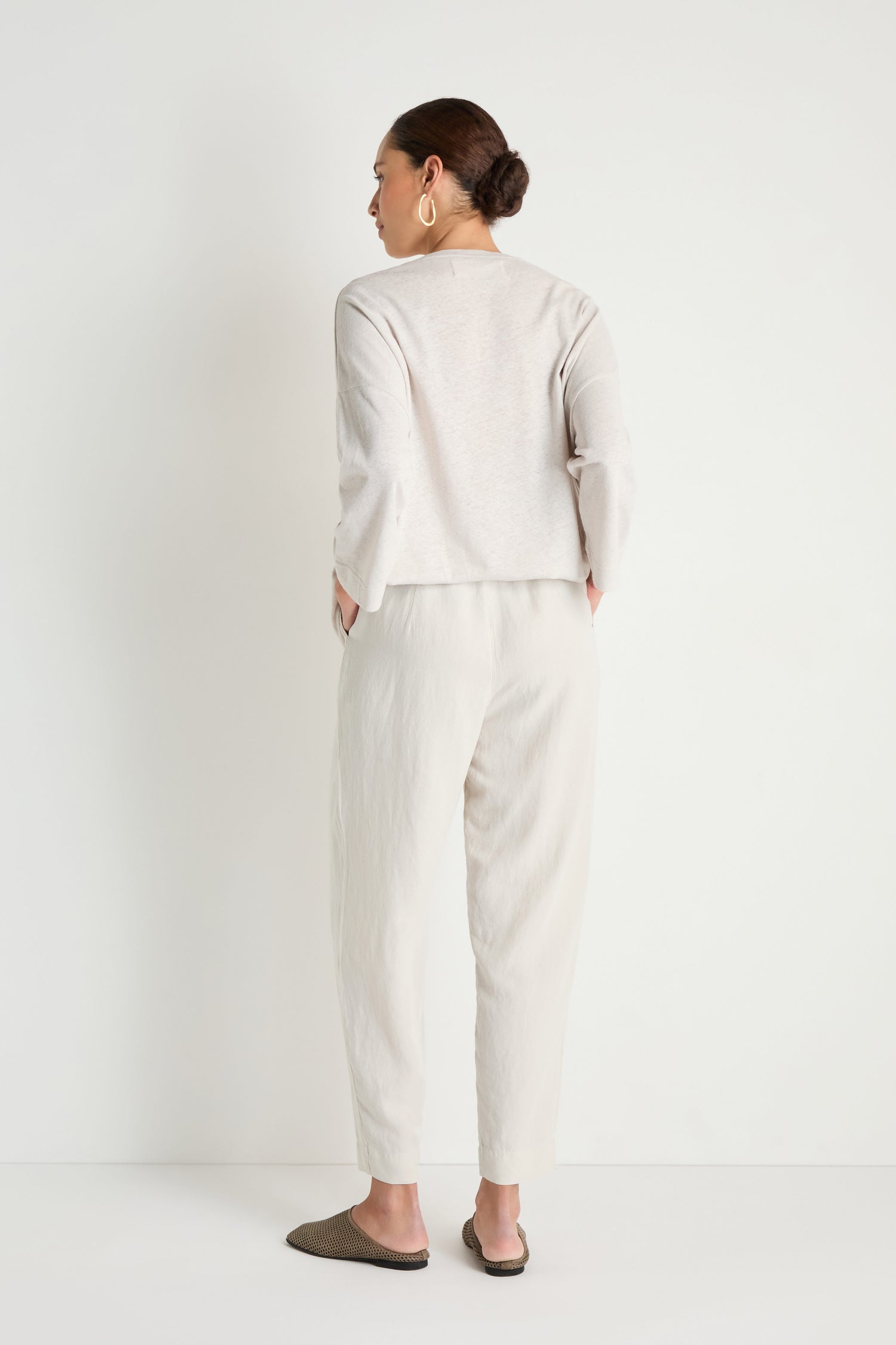 A woman faces away from the camera against a white background, wearing a light long-sleeve top, Cream Lyocell Linen Slim Trousers with an elasticated waist, hoop earrings, and flat shoes.