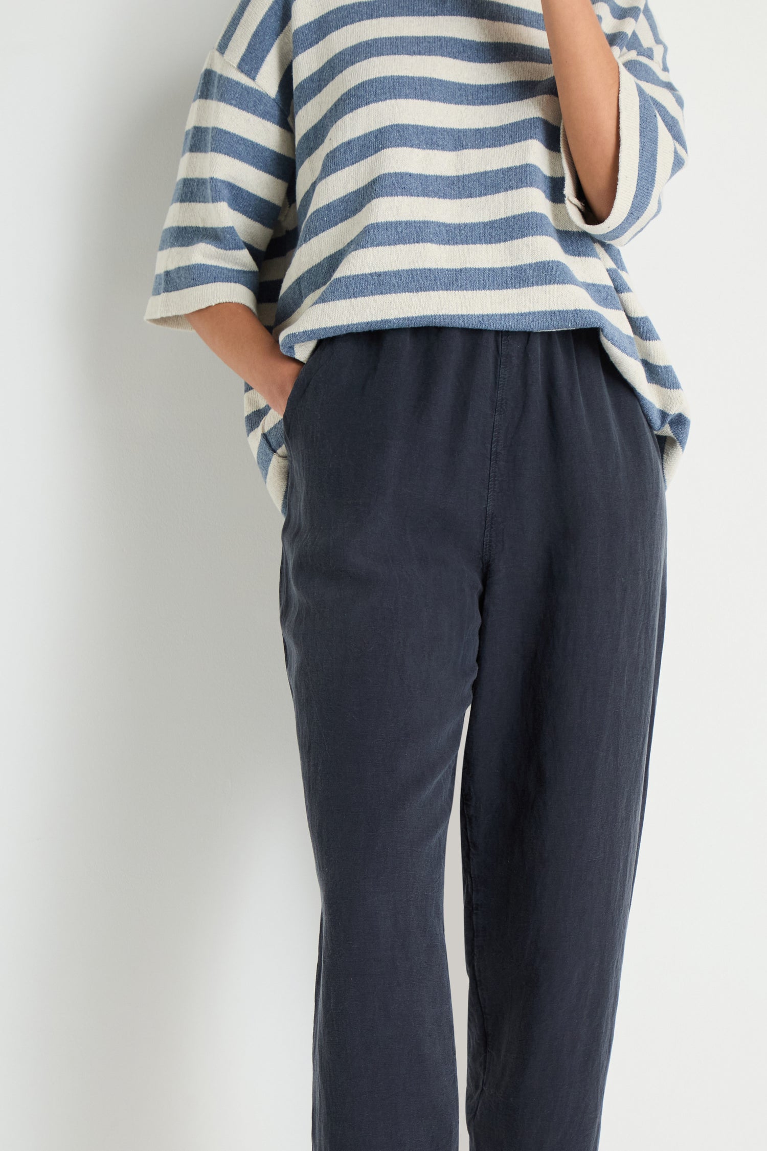 Person wears Navy Lyocell Linen Slim Trousers with a blue and white striped top, standing against a plain light background. Face not visible.