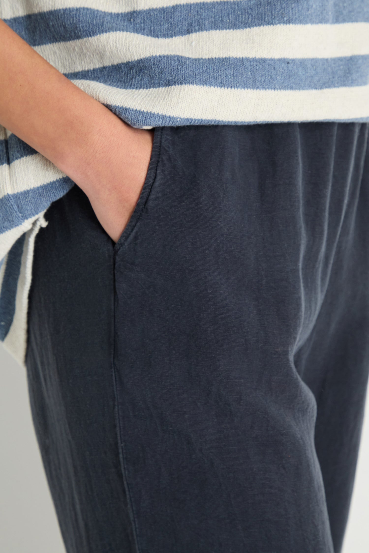A person stands with one hand in the pocket of Navy Lyocell Linen Slim Trousers, paired with a blue and white striped top.