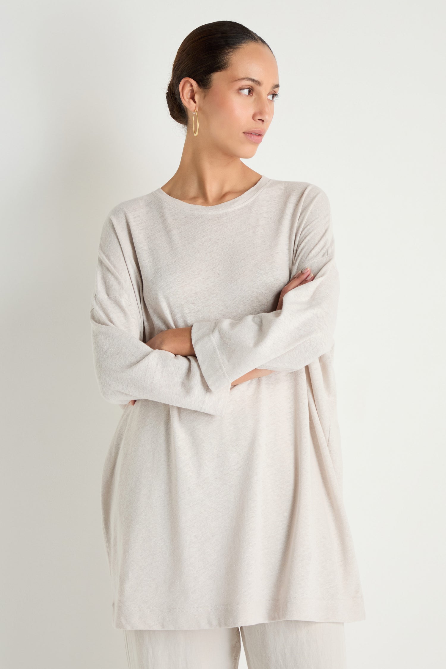 A woman stands with arms crossed, wearing the Linen Cotton Jersey Tunic in light beige with matching pants, showcasing a relaxed silhouette against a plain background.