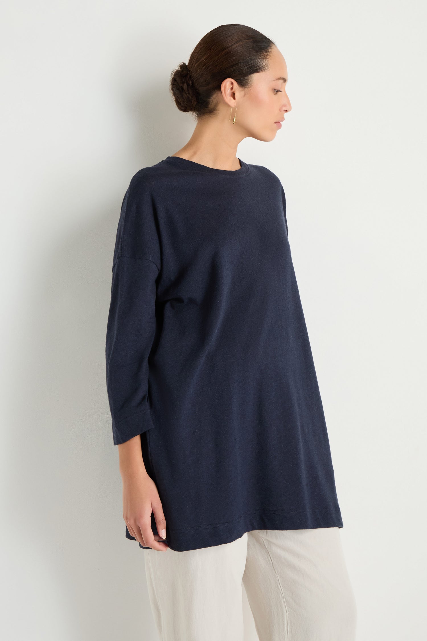 A woman with her hair in a bun stands sideways against a white wall, wearing the Linen Cotton Jersey Tunic in navy with a relaxed silhouette, paired with white pants.
