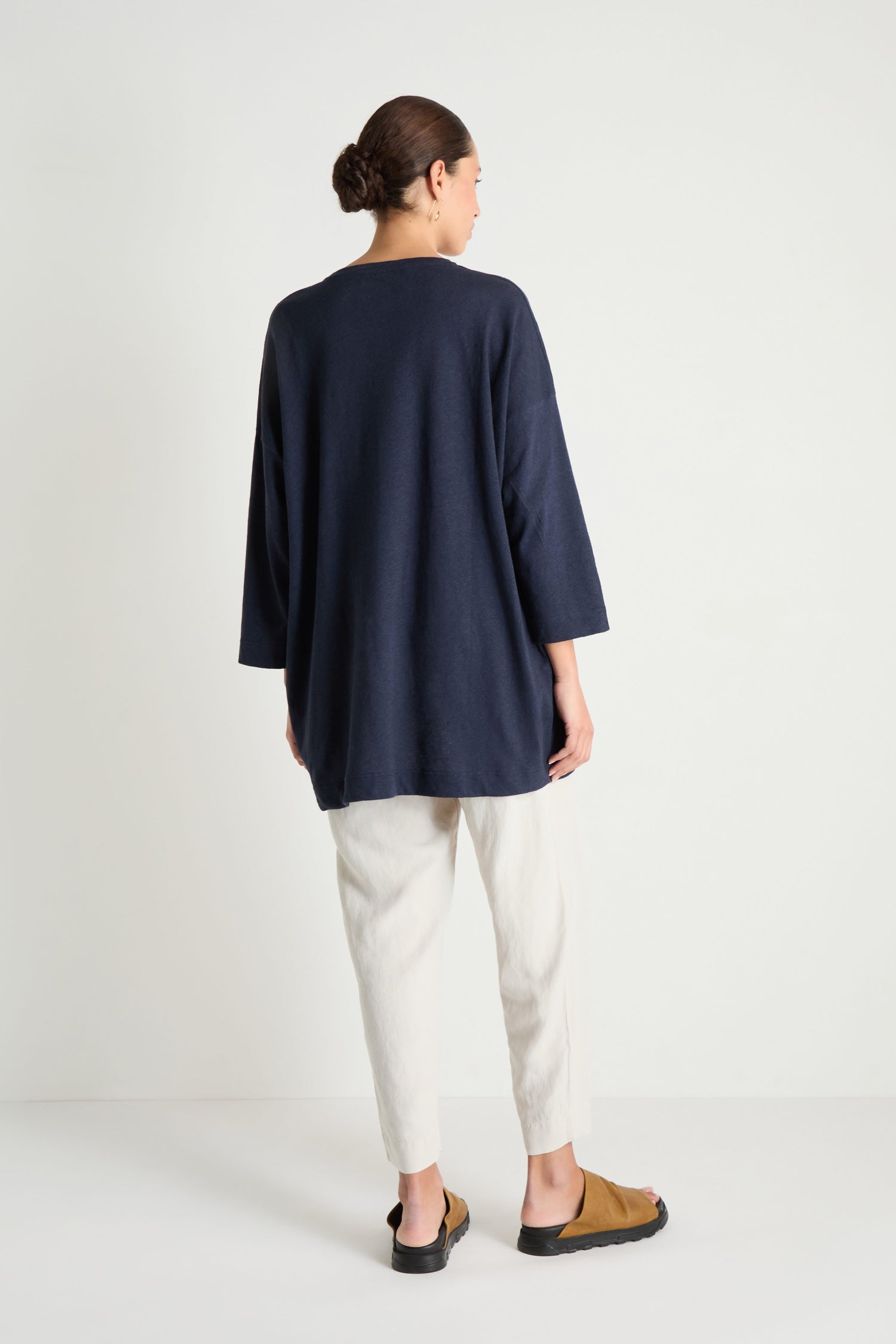 A person stands facing away from the camera in a Linen Cotton Jersey Tunic, featuring a relaxed silhouette, paired with light pants and brown sandals, set against a plain white background.