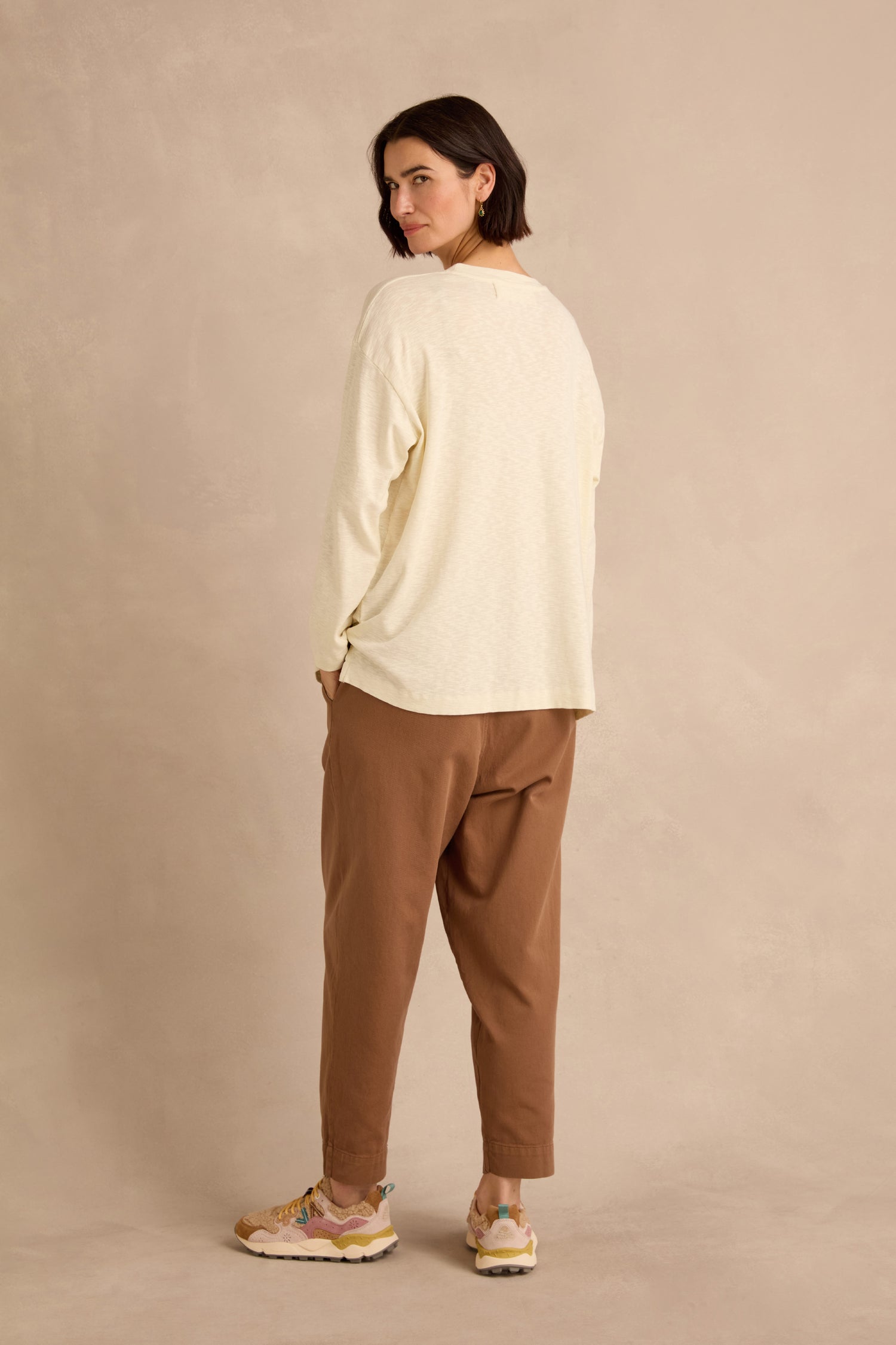 A woman with dark hair is wearing the Cream Boxy Cotton Jersey T Shirt—featuring long sleeves and a loose, longline fit—paired with brown pants and sneakers as she stands facing away and looks over her shoulder against a beige background.