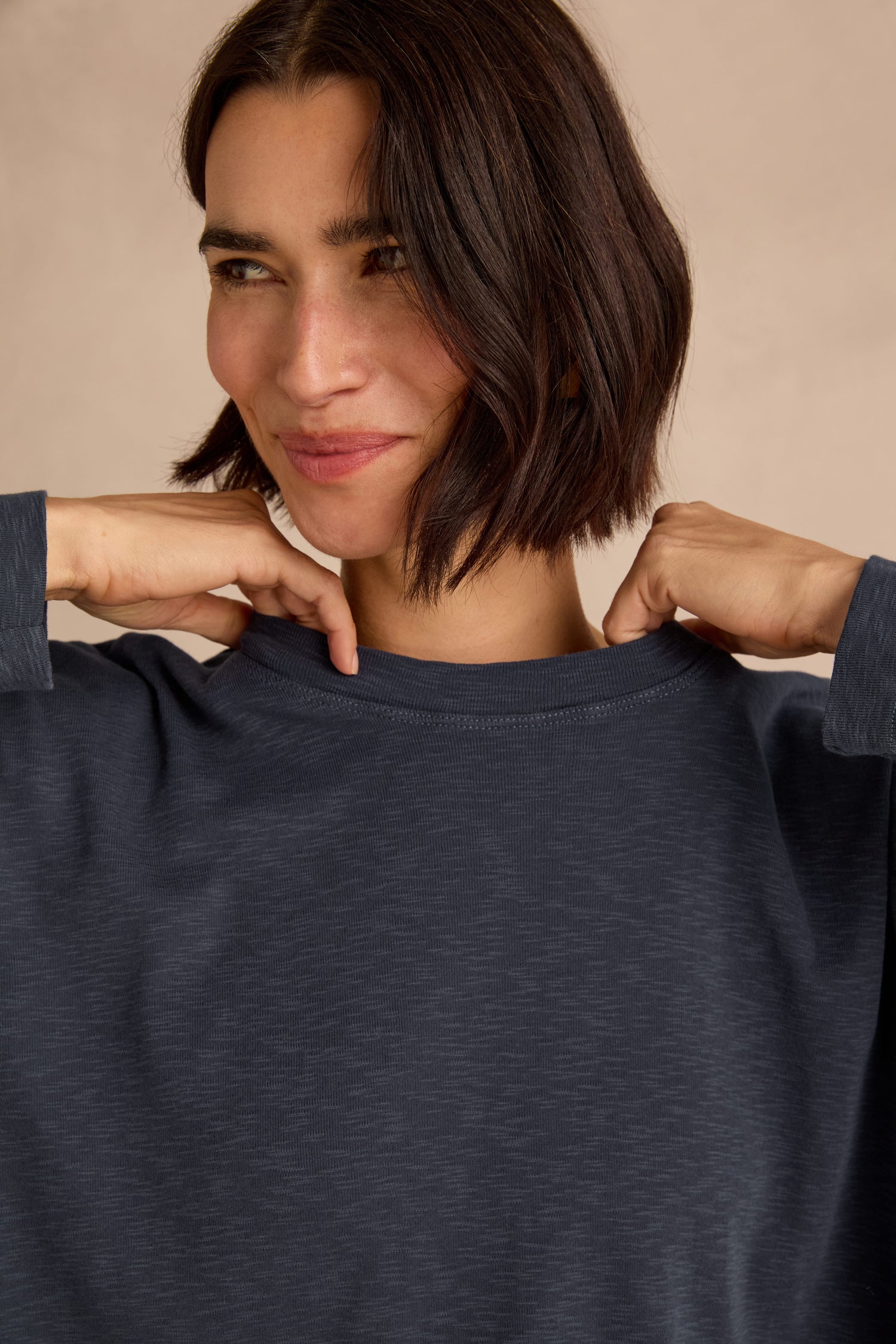 A woman with short dark hair wears the Navy Boxy Cotton Jersey T Shirt in a loose, longline fit. She smiles slightly, raising her hands near her collar against a neutral background.