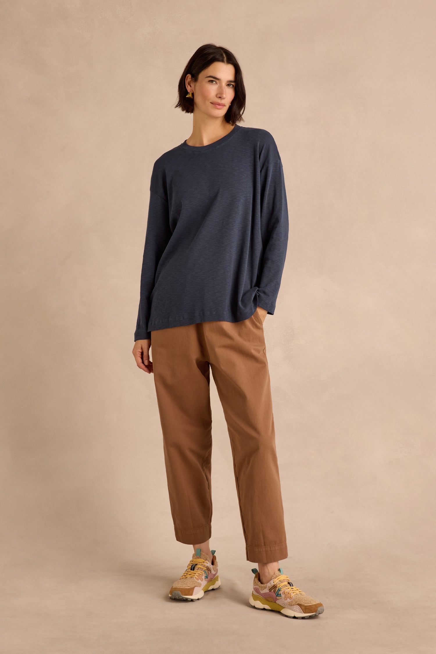 A woman stands against a beige backdrop wearing the Navy Boxy Cotton Jersey T Shirt with a loose, longline fit, paired with light brown pants and yellow sneakers.