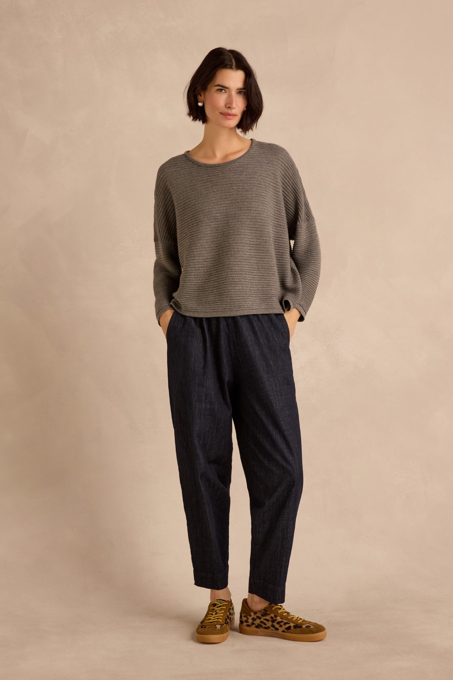Wearing the Mole Fine Rib Merino Top in grey, a person stands against a beige background with hands in their pockets, paired with dark blue pants and leopard print sneakers.
