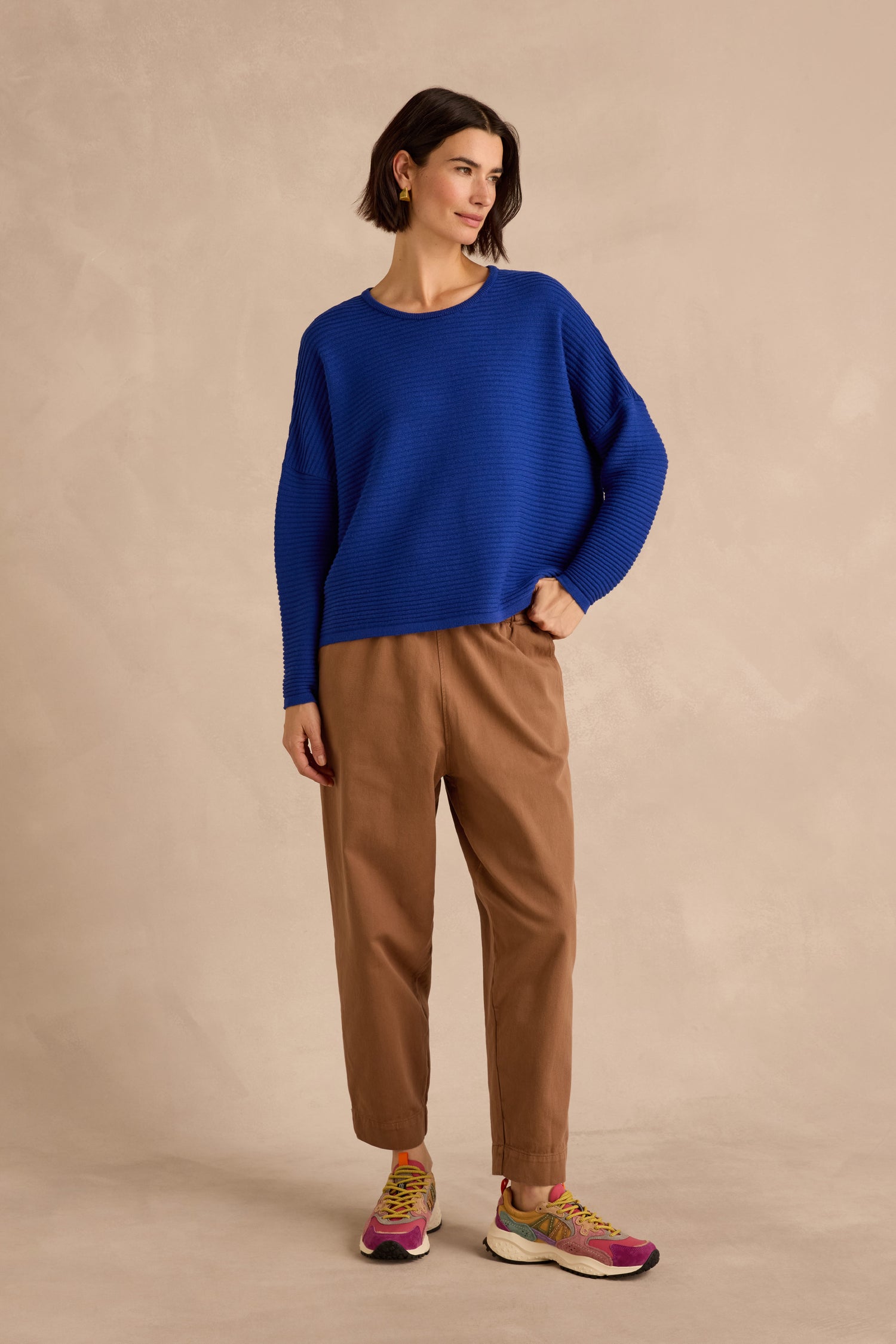 A person stands against a beige background, wearing the Royal Blue Fine Rib Merino Top in a relaxed boxy fit, paired with tan pants and colorful sneakers, with one hand in their pocket.