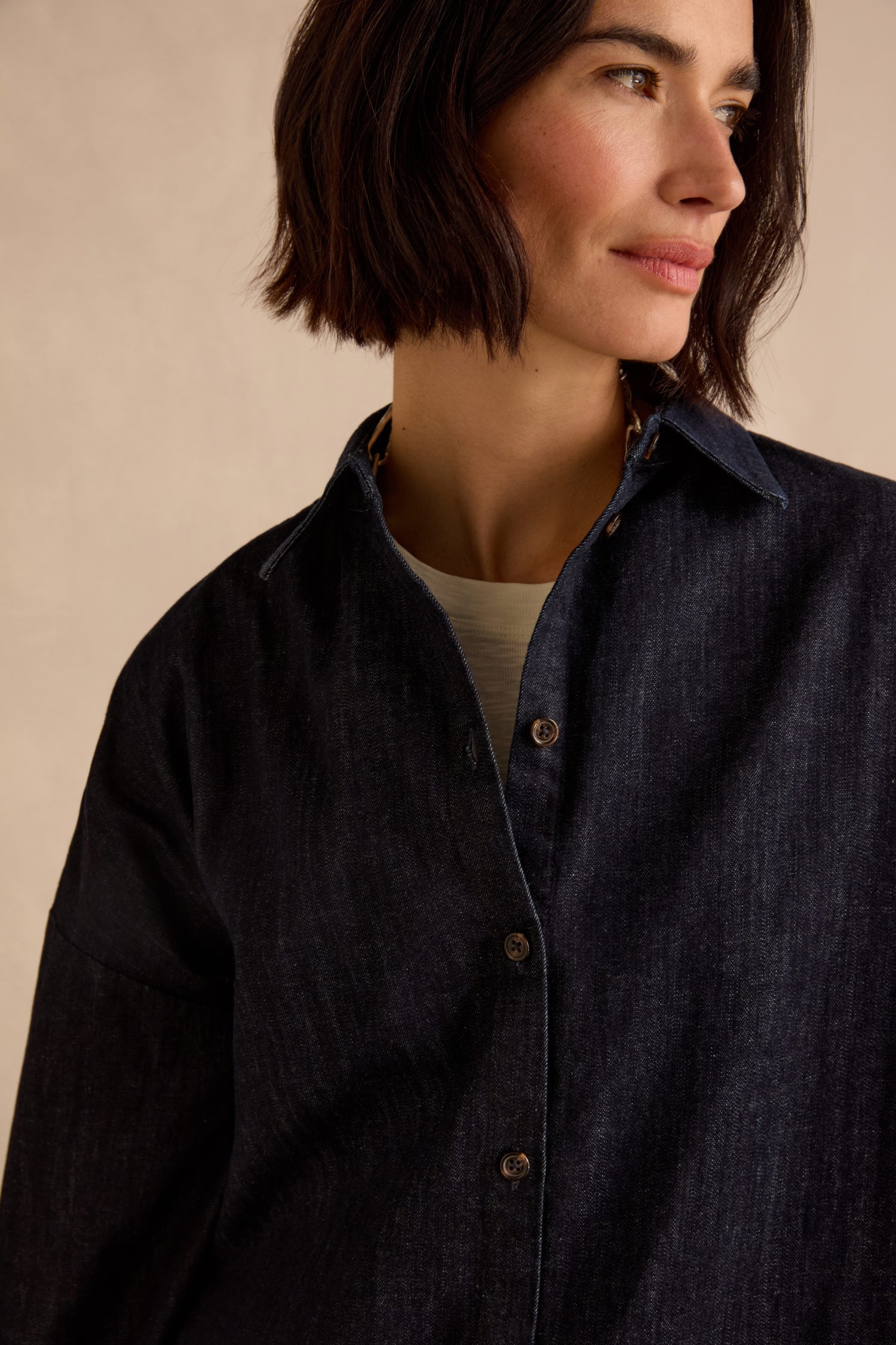 A woman with short dark hair models the Denim Oversized Shirt, crafted from pure cotton in an indigo wash, layered over a light top, as she looks to the side against a neutral background.