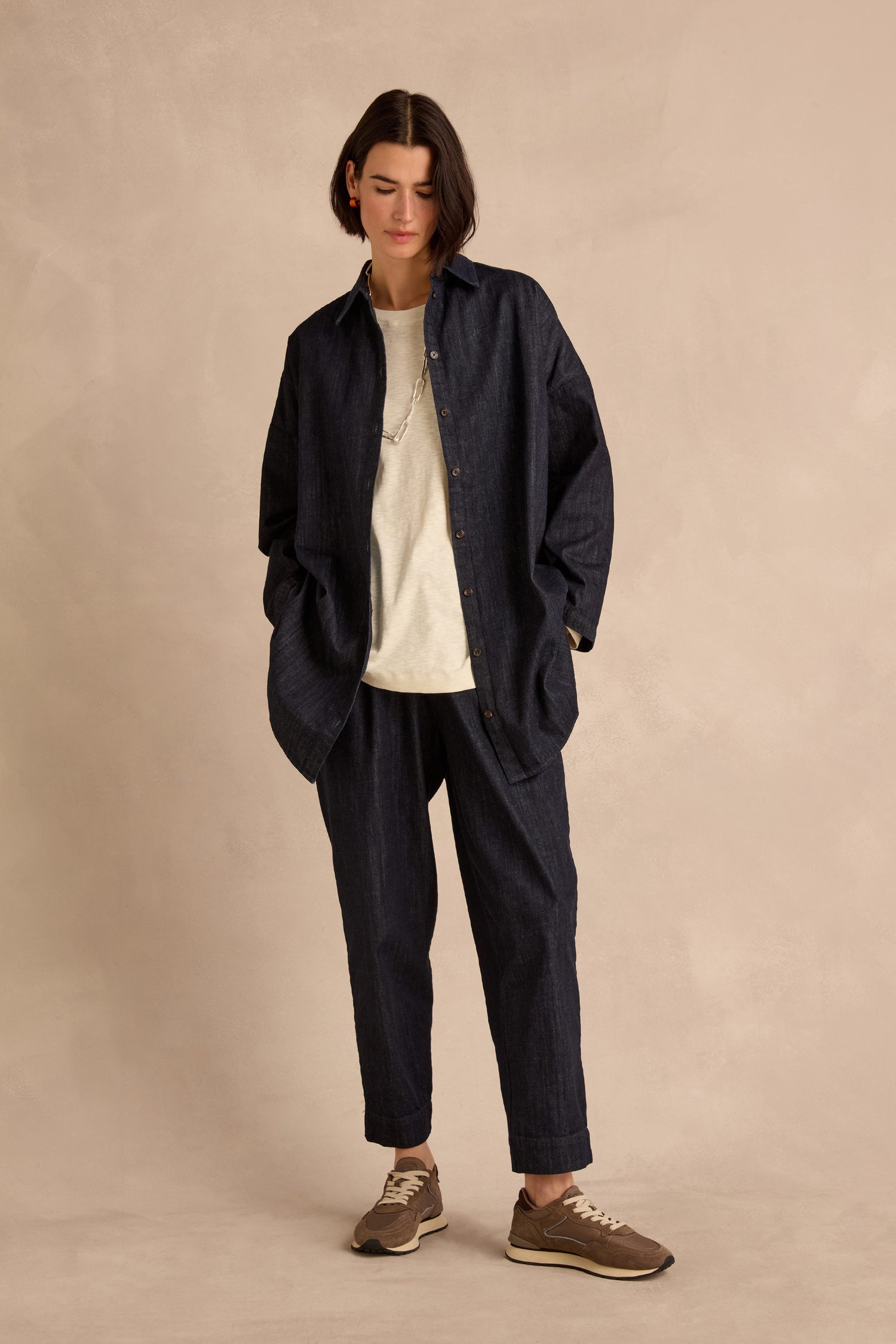 A person is wearing the Denim Oversized Shirt in an indigo wash, crafted from pure cotton denim, styled with matching pants over a white t-shirt, brown sneakers, and a chain necklace, standing against a beige background.
