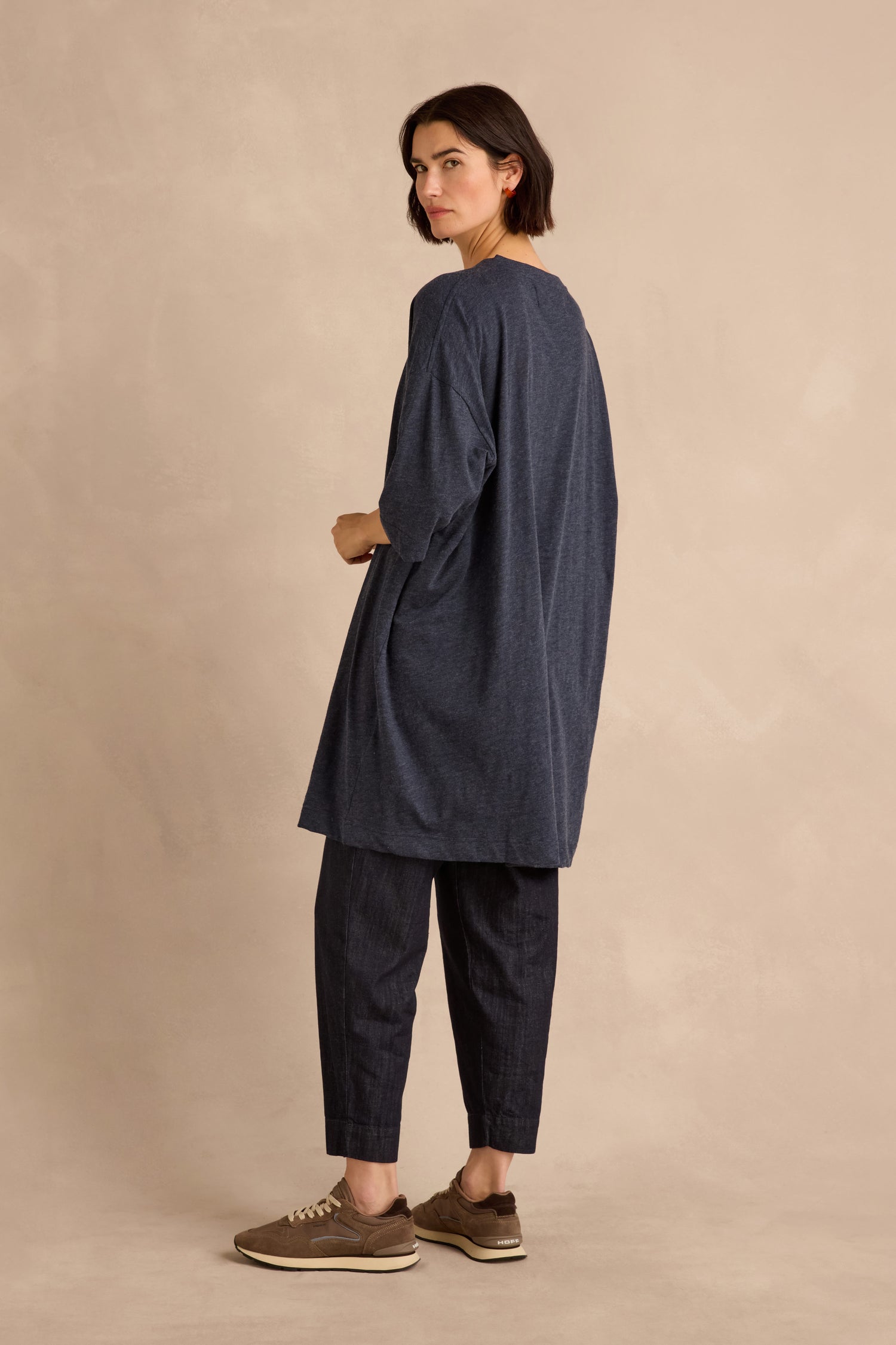 A woman stands sideways against a beige background, wearing the Cotton Wool Jersey Tunic in navy with a relaxed silhouette, matching pants, and brown sneakers.