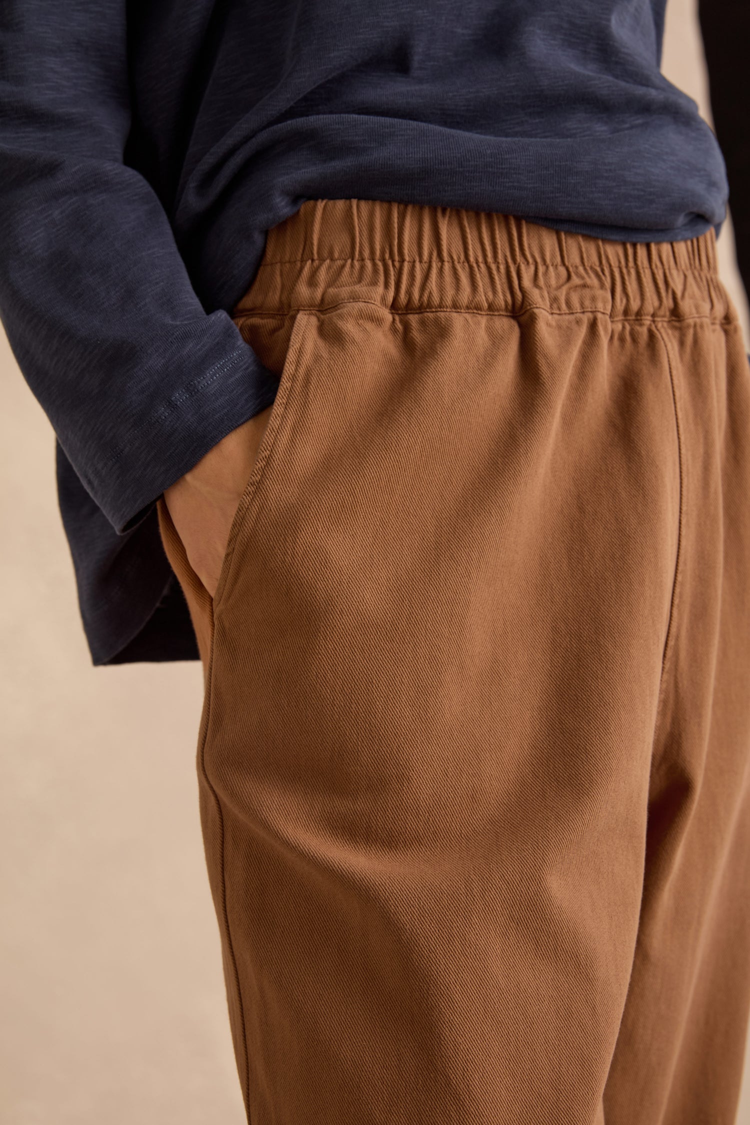 Person wearing a navy long-sleeve shirt and Camel Laura Tapered Denim Trousers with an elasticated waistband, their hand in the pocket.