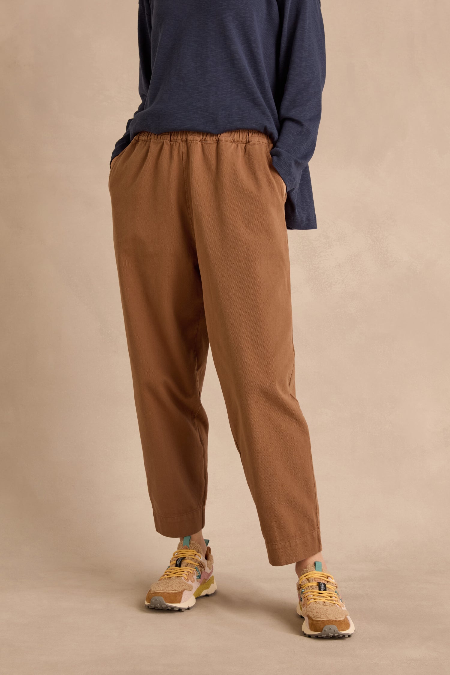 A person wearing a navy long-sleeve top, Camel Laura Tapered Denim Trousers with an elasticated waistband, and yellow sneakers stands with hands in pockets against a beige background.
