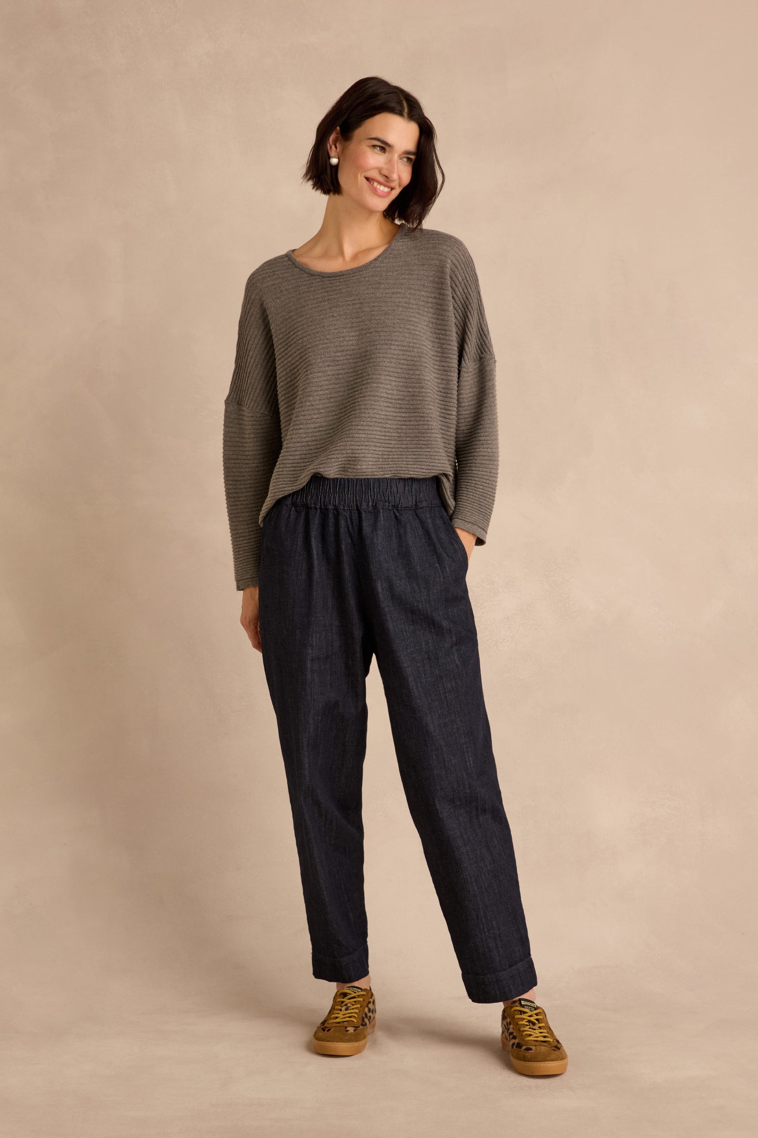 A woman stands against a beige background, smiling and looking slightly to the side. She is wearing a loose gray sweater, brown sneakers, and the Laura Tapered Denim Trousers with an elasticated waistband in dark blue.