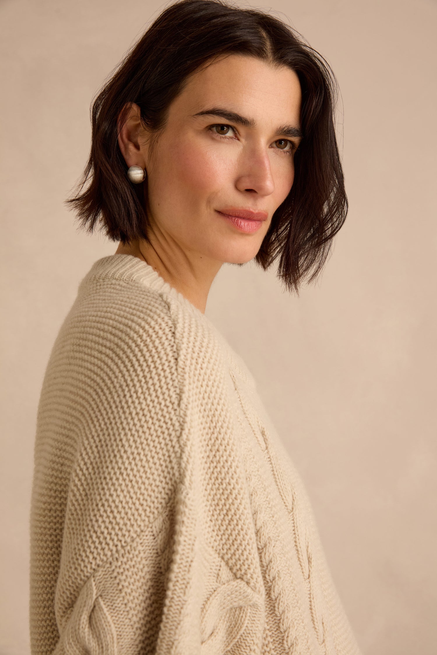 Woman with short dark hair wearing the Cable Detail Wool Tunic in a cream recycled wool knit, styled with pearl earrings, standing against a neutral background.