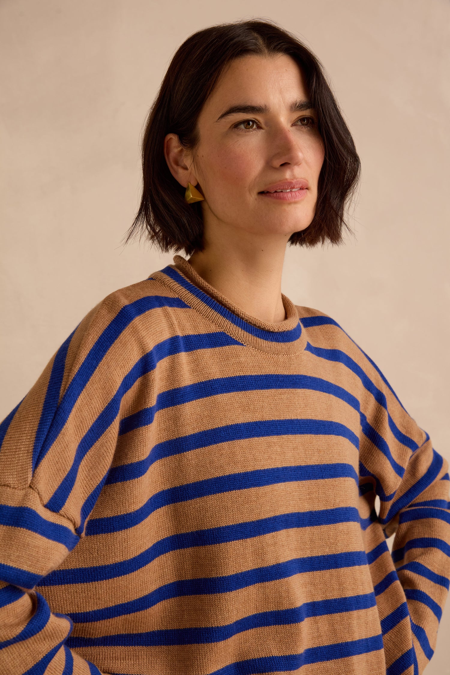 A woman with short dark hair wears the Royal Blue Stripe Boxy Merino Knit, a brown and blue striped sweater crafted from soft merino wool. She stands against a beige background and gazes slightly to the side.