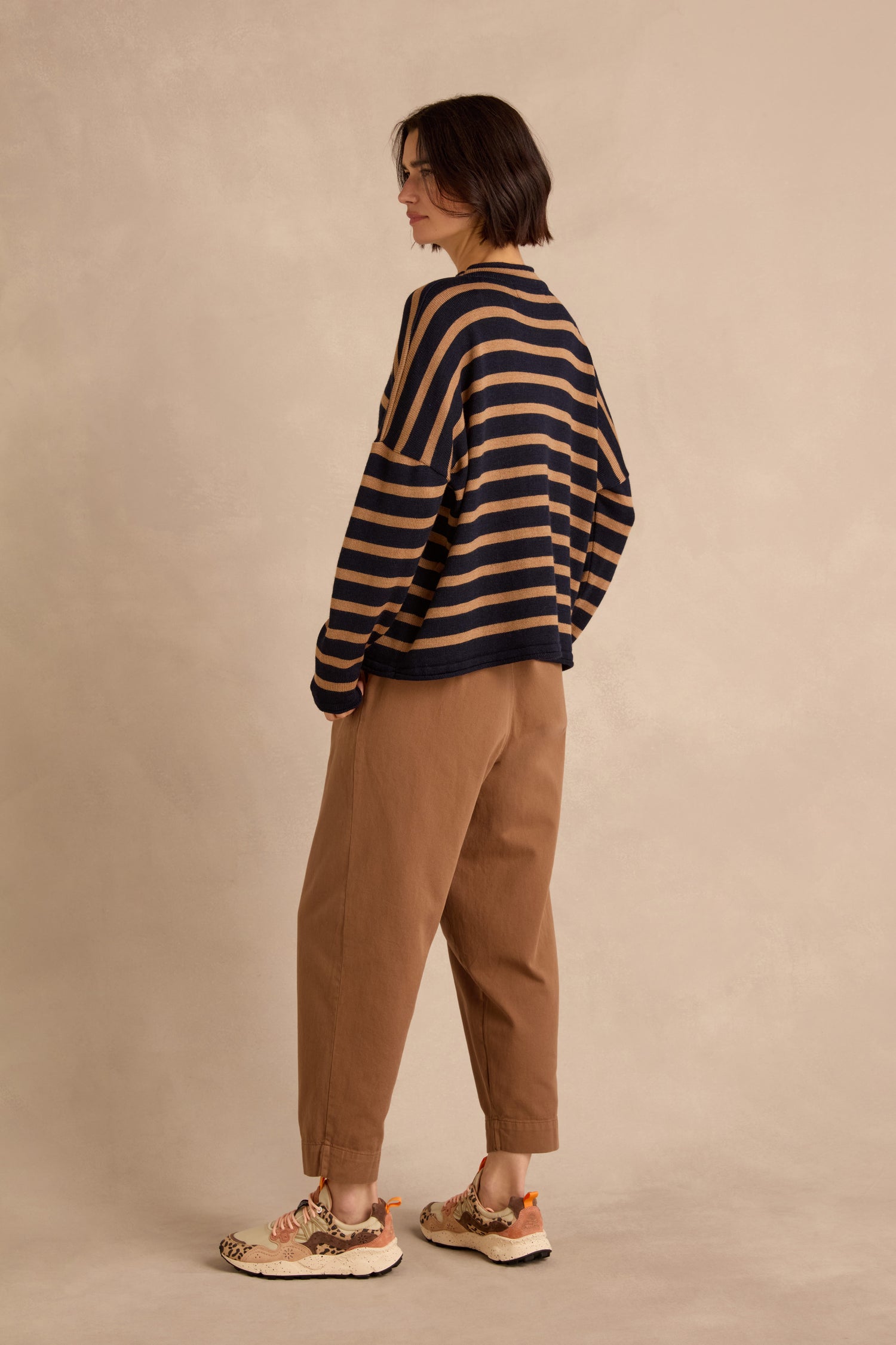 Wearing the Navy Stripe Boxy Merino Knit, a soft merino wool sweater featuring navy and tan stripes, paired with brown pants and sneakers, the person stands sideways against a plain beige background.