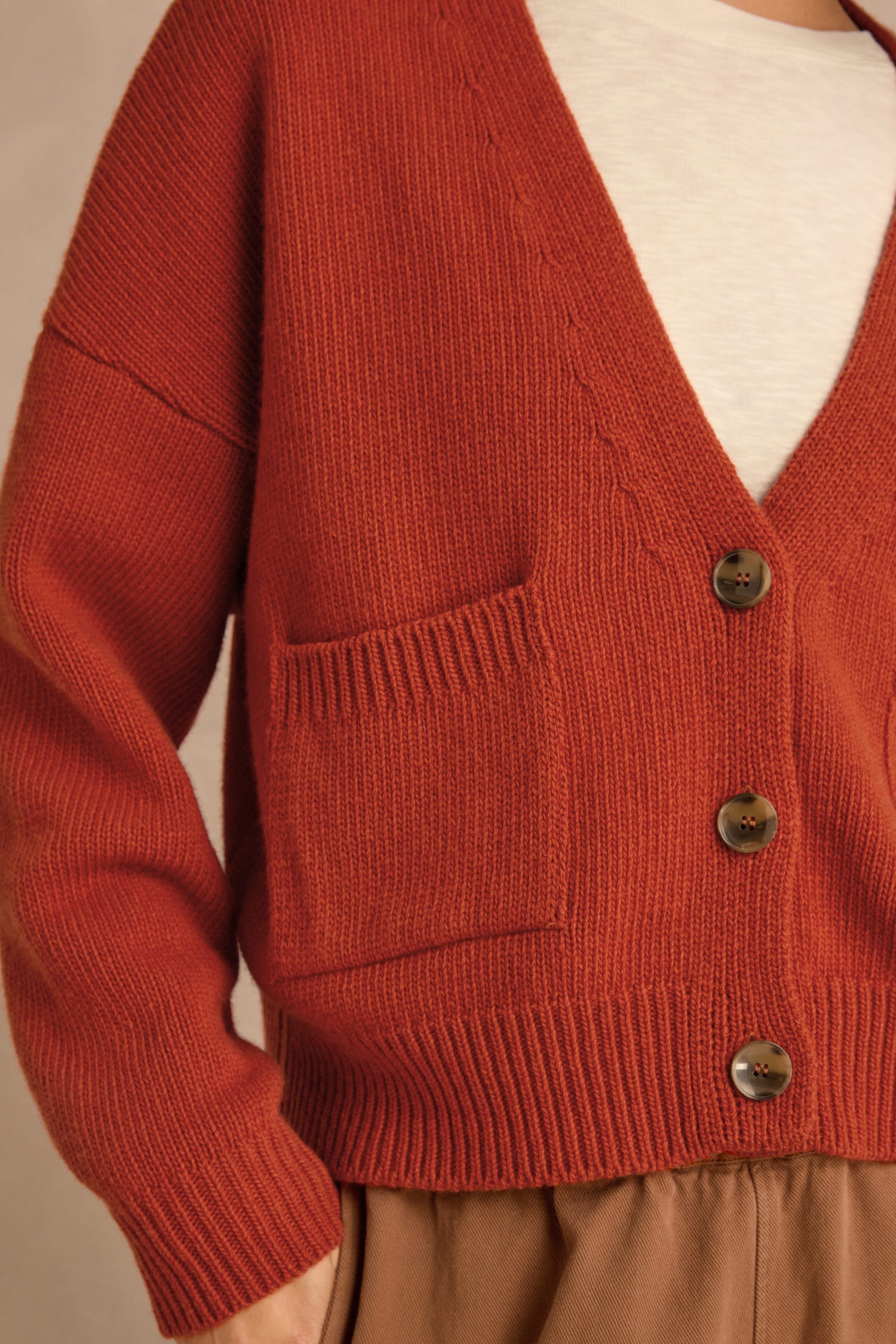 A person wearing the Wool Boxy Cardigan in rust, featuring brown buttons and a front pocket, styled over a white top and tan pants.