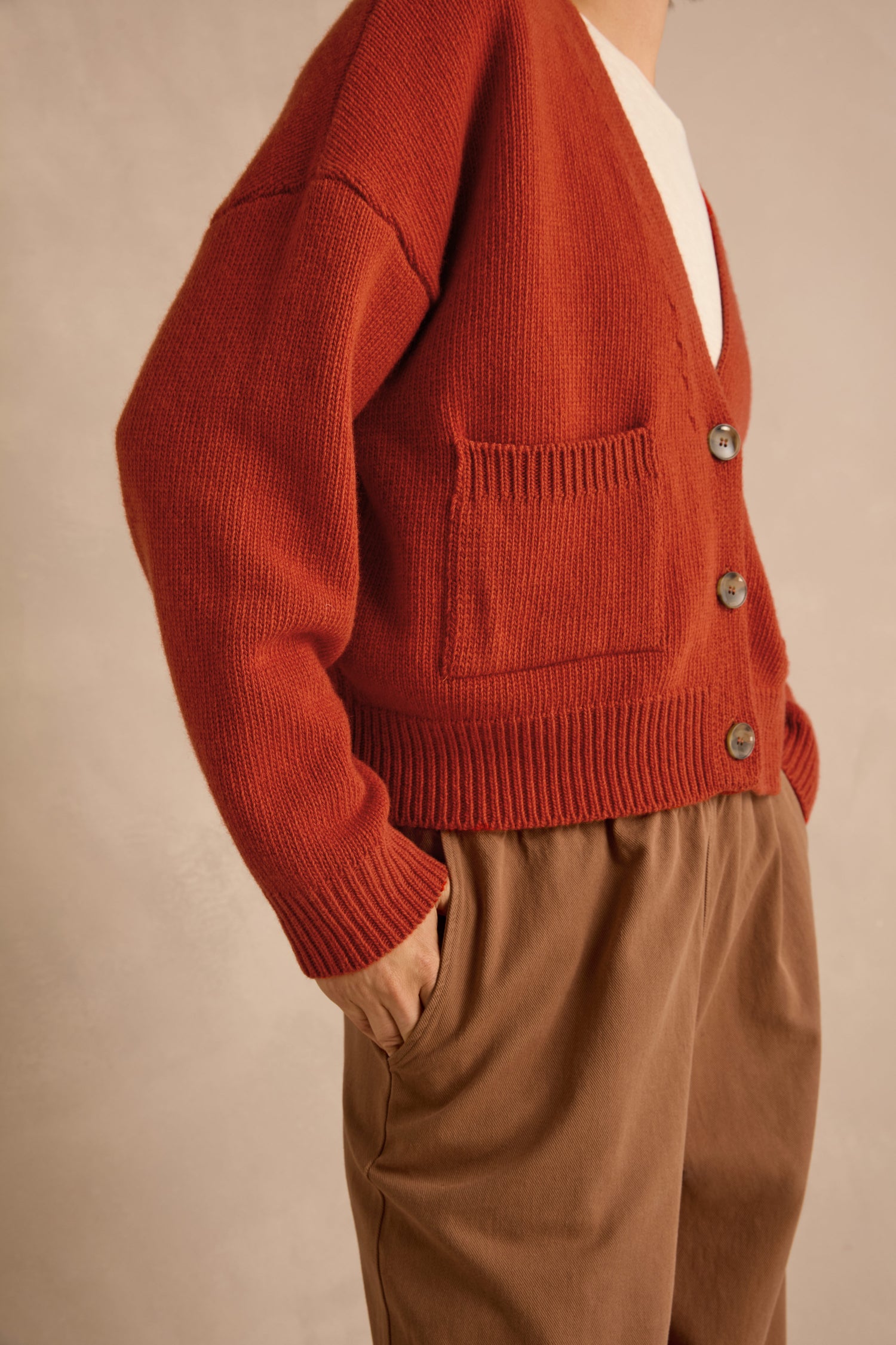 A person wearing the Wool Boxy Cardigan in a rust color and brown pants stands with one hand in their pocket against a neutral background.