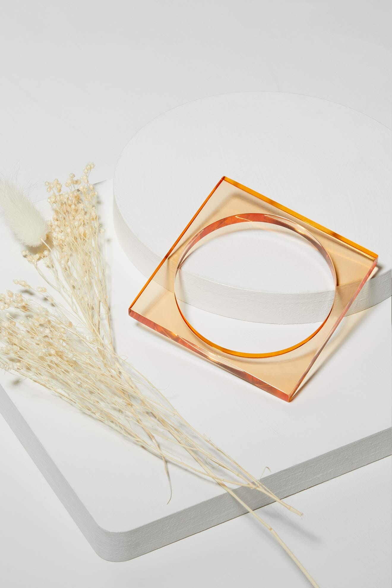 Square Bangle: a translucent orange modern accessory displayed on white geometric platforms next to dried, light-colored flowers.