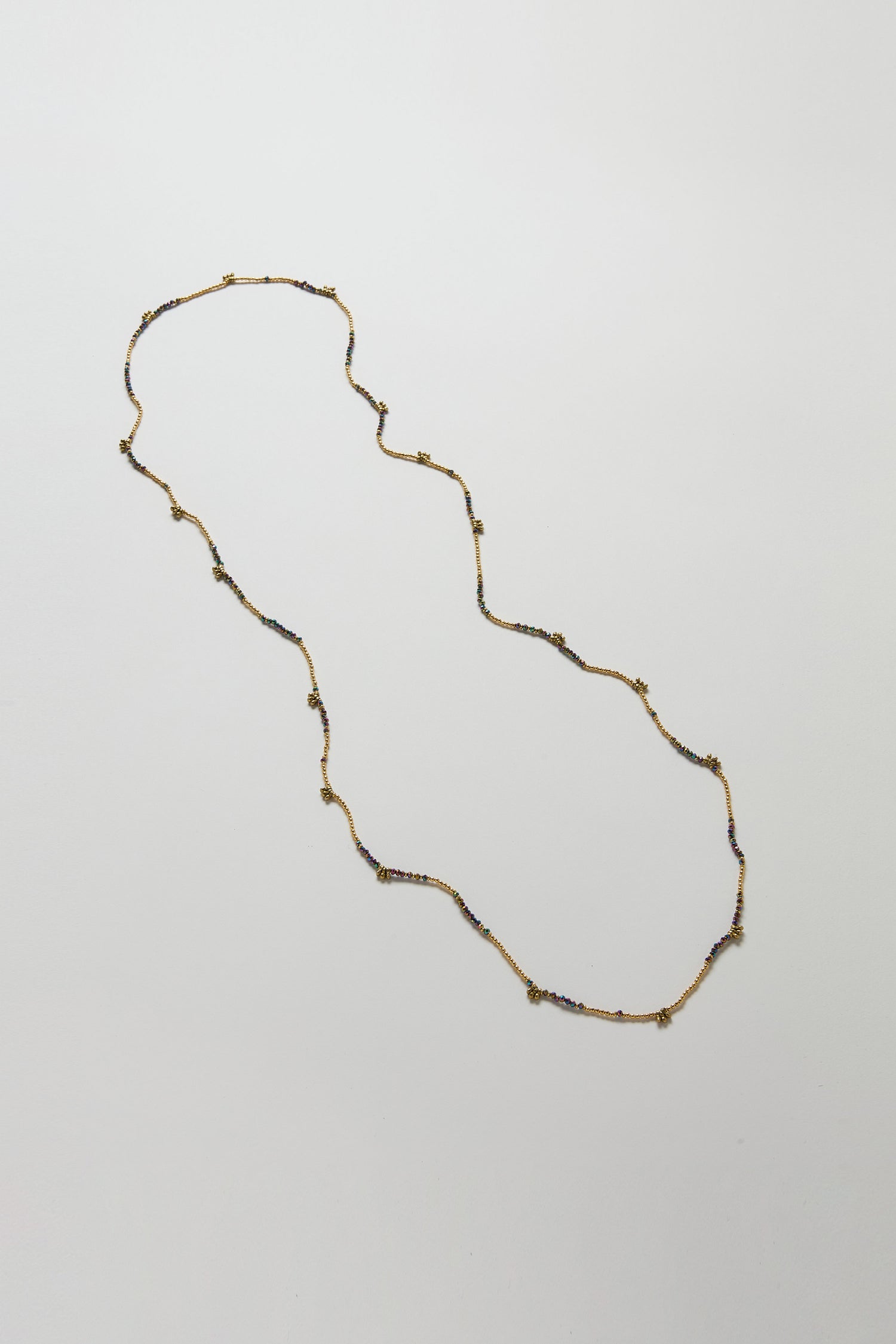 The Long Saros Beaded Necklace showcases a slender gold chain adorned with evenly spaced beads, elegantly displayed in a gentle curve against a plain white background.