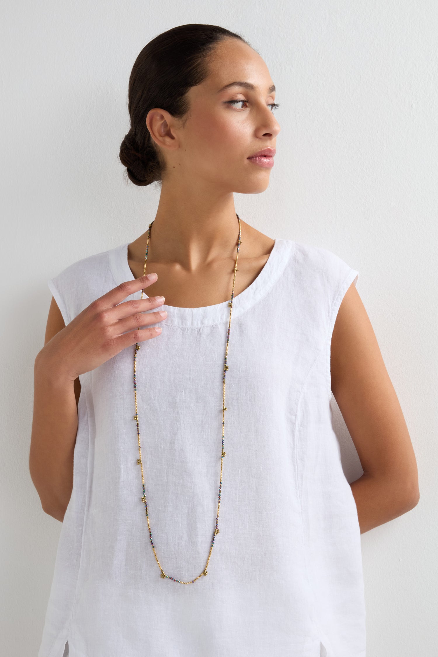 Woman wearing a white sleeveless top and the Long Saros Beaded Necklace, crafted with multicoloured seed beads, standing against a plain white wall and looking to the side.
