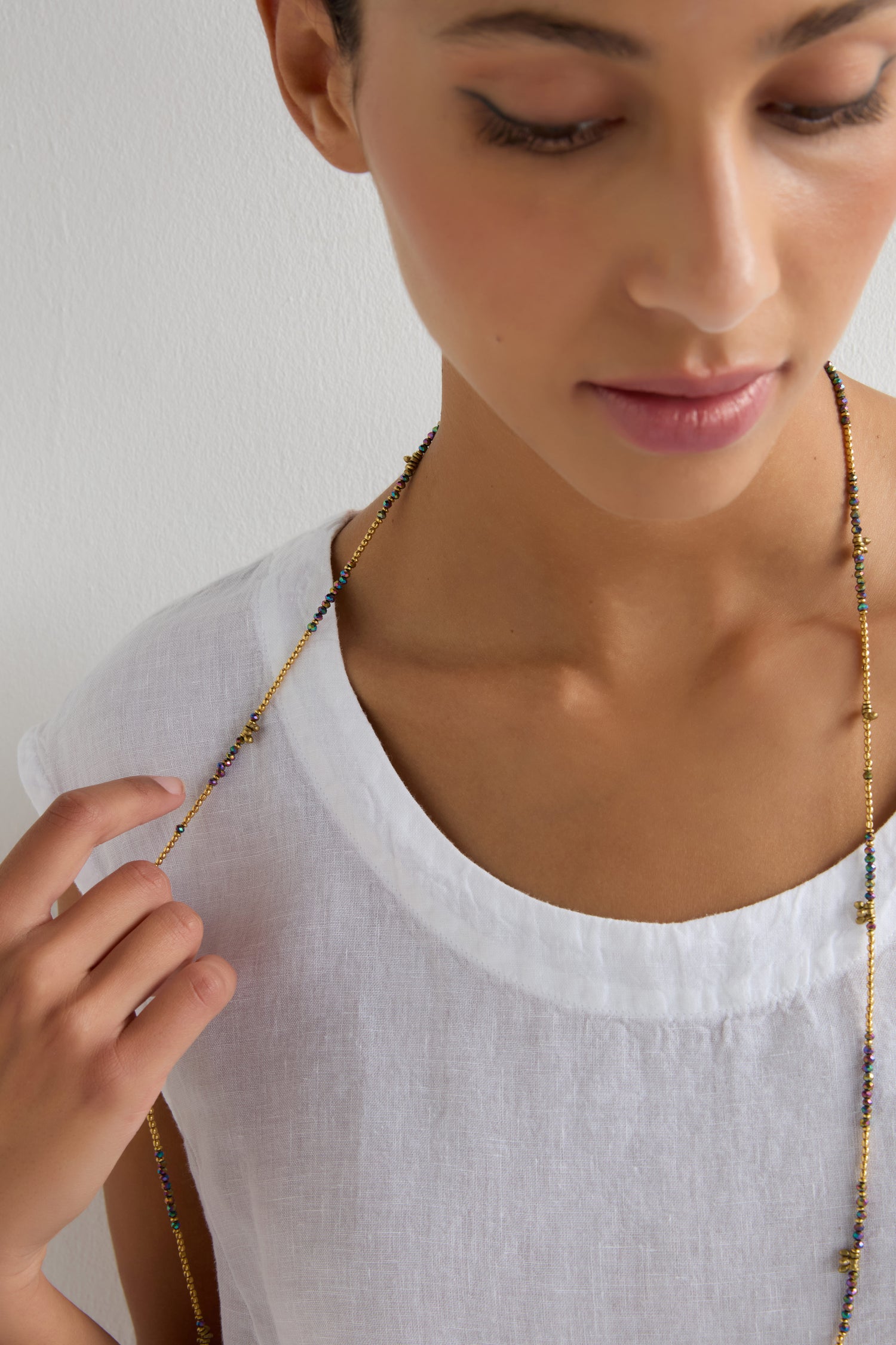 A person in a white linen top holds the Long Saros Beaded Necklace, its multicoloured seed beads from the Nataraj Collection resting near their chest as they gaze downward with a neutral expression.