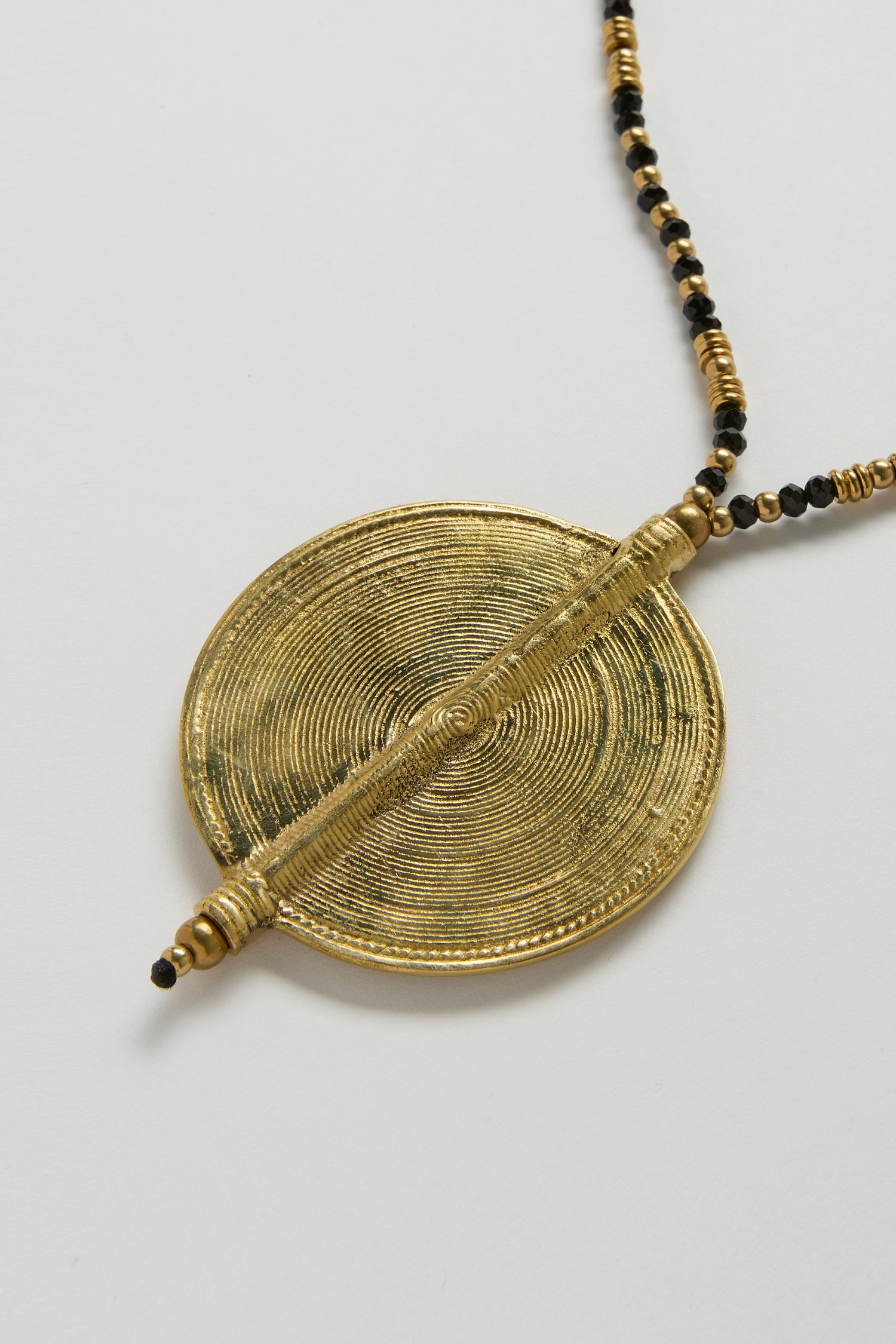 The Long Spiral Pendant, from the Nataraj Collection, showcases concentric ridges on a circular gold and black design, paired with a beaded spiral necklace and presented on a white background.