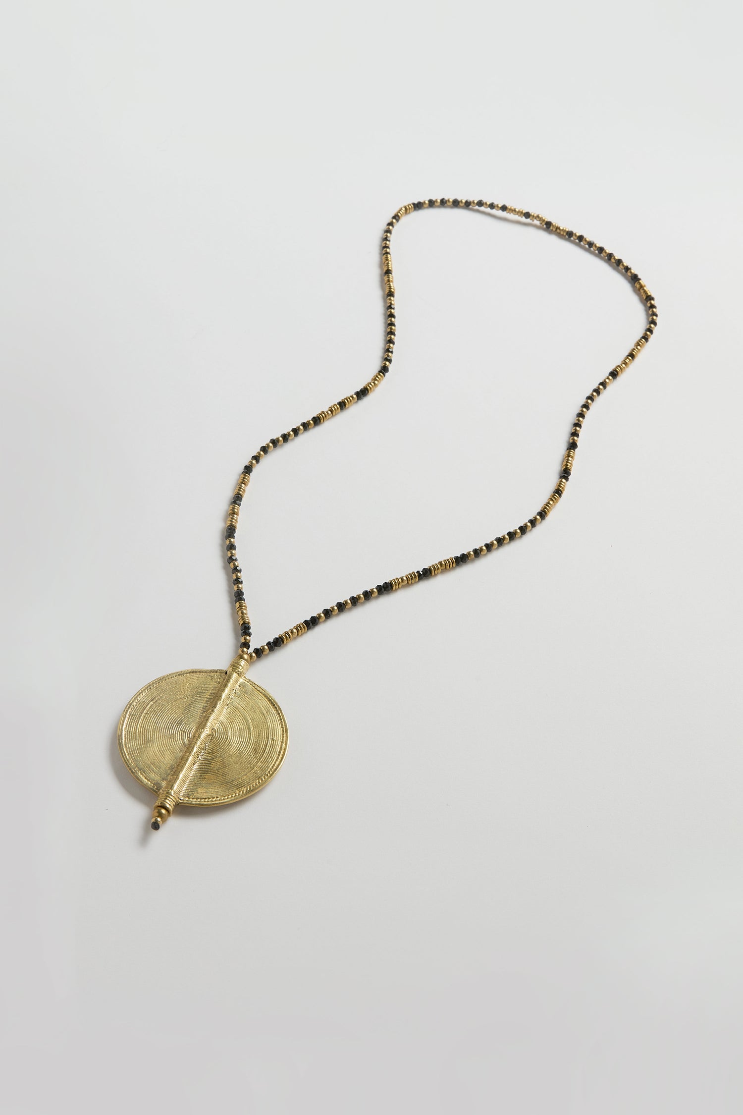 The Long Spiral Pendant, part of the Nataraj Collection, features a beaded necklace with a large round gold embossed pendant and is displayed on a plain white background.