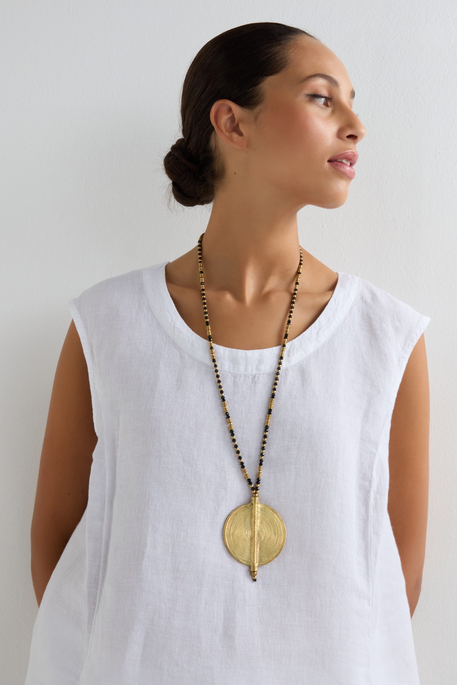 A woman in a sleeveless white top models the Long Spiral Pendant, featuring gold and black tones and artisanal craftsmanship, as she looks to her right against a plain white background.