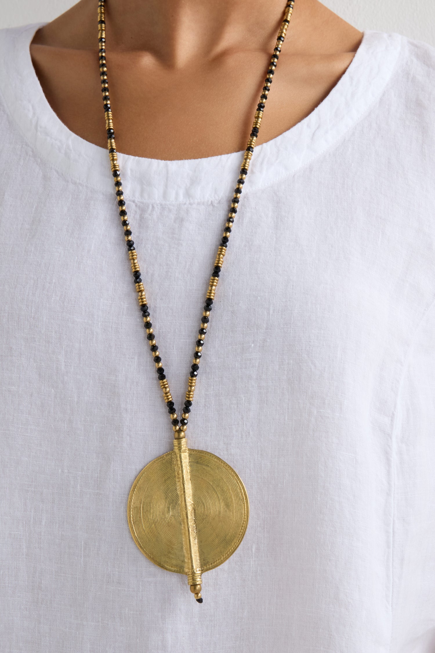 A person wearing a white linen shirt with a beaded necklace showcasing the Long Spiral Pendant in gold and black, a statement piece of artisanal craftsmanship.