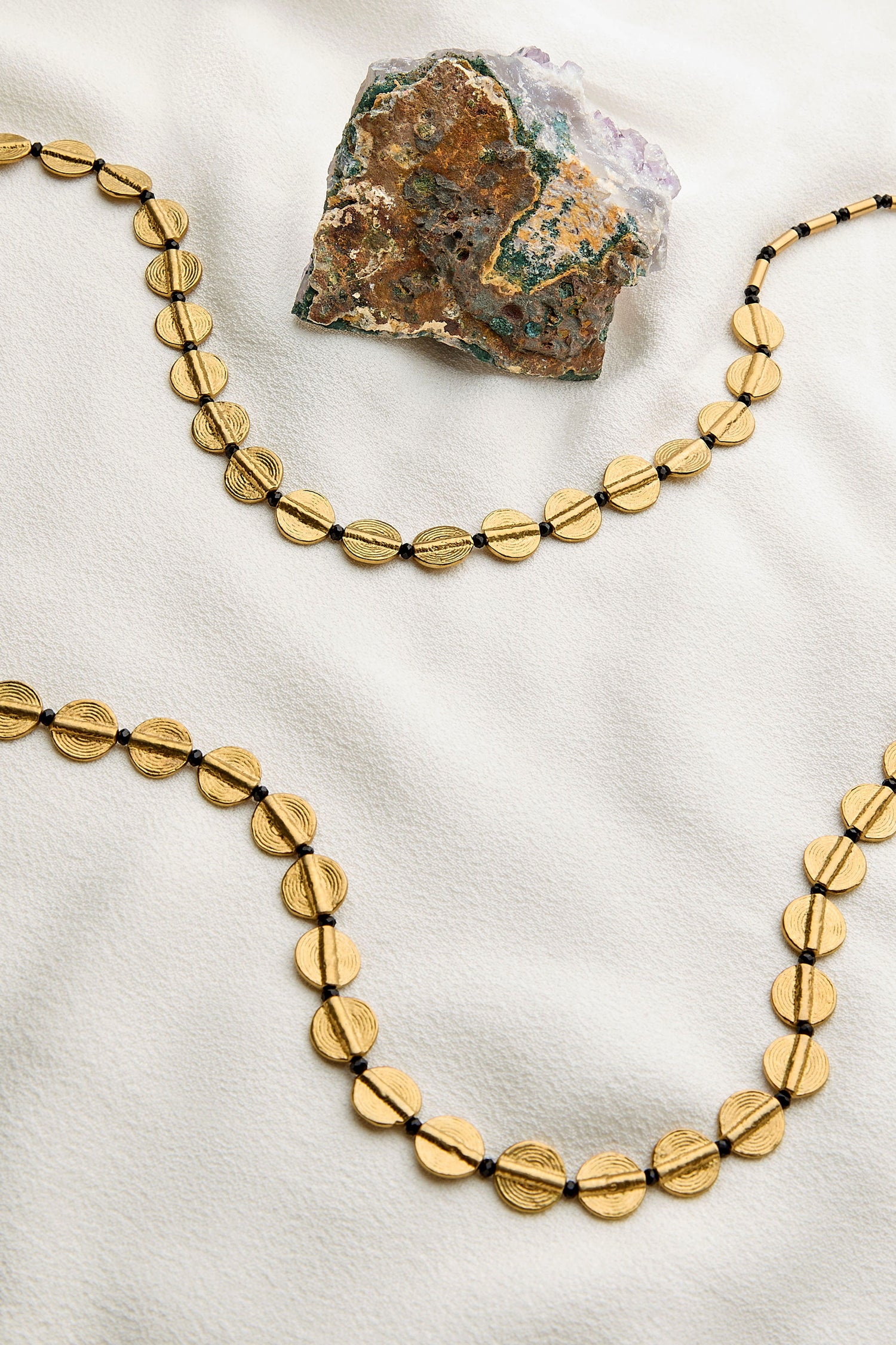 The Long Engraved Circles Necklace, handcrafted by Jaipur artisans, displays round engraved circles arranged in curves on white fabric beside a rough, multicolored stone.