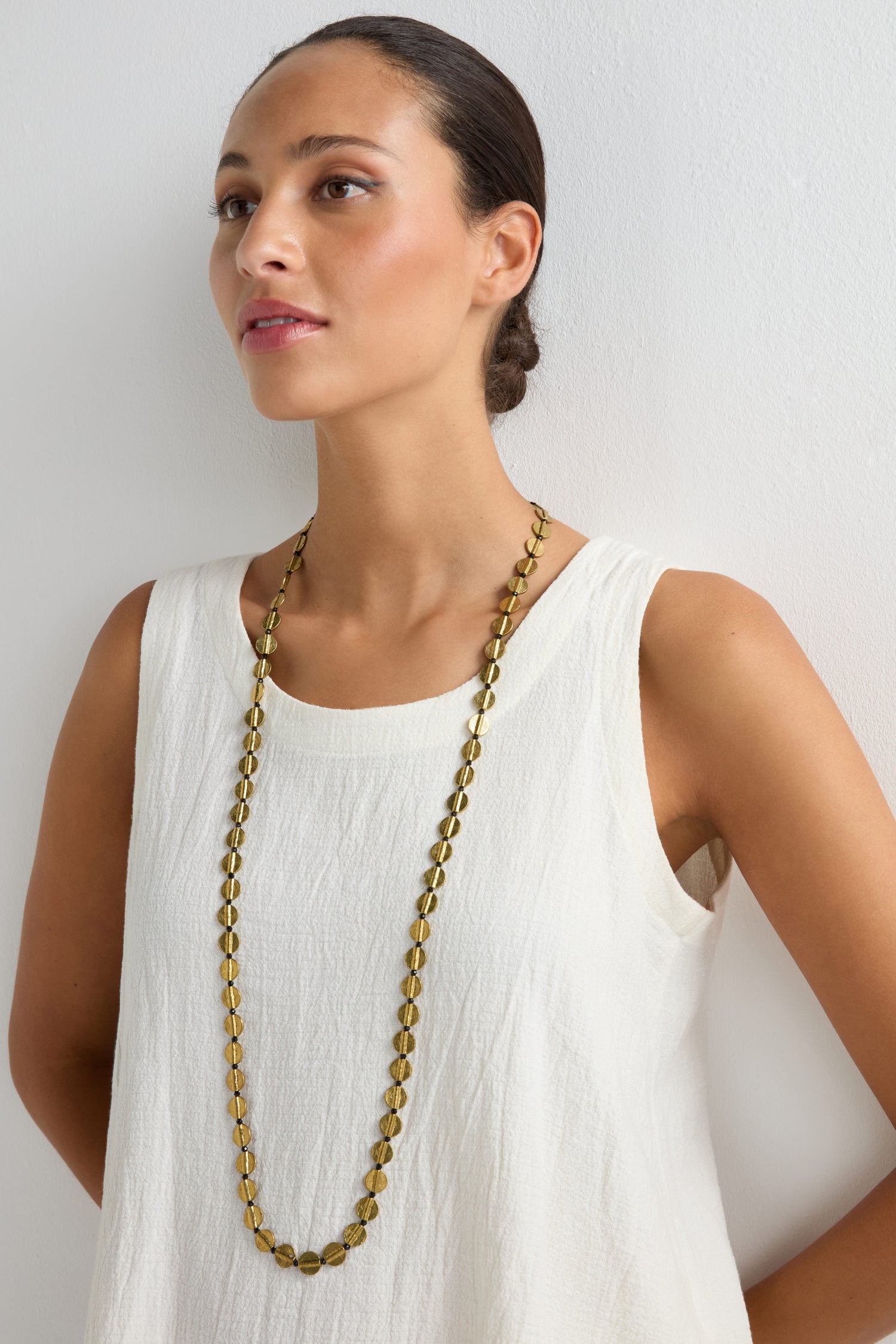 A woman in a sleeveless white top wears the Long Engraved Circles Necklace, handcrafted by Jaipur artisans, standing against a plain white background.