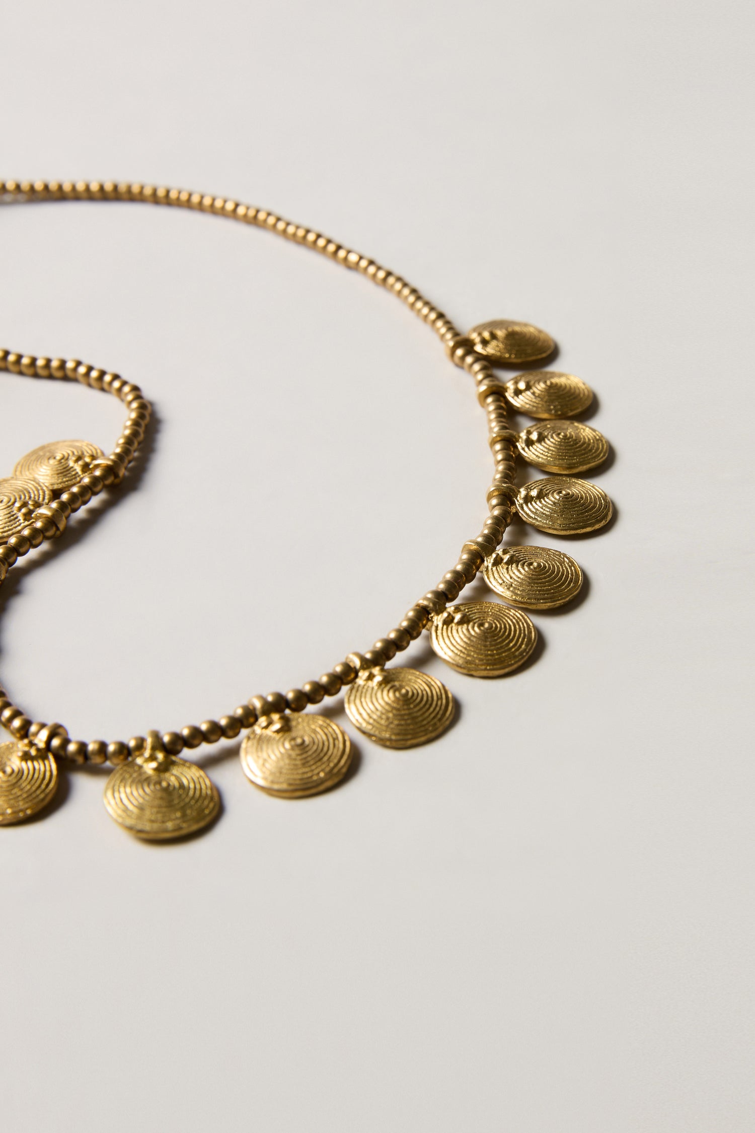 The Short Gold Chakra Necklace features evenly spaced, textured golden discs as pendants on a chain, shown against a plain light background—a timeless statement piece.