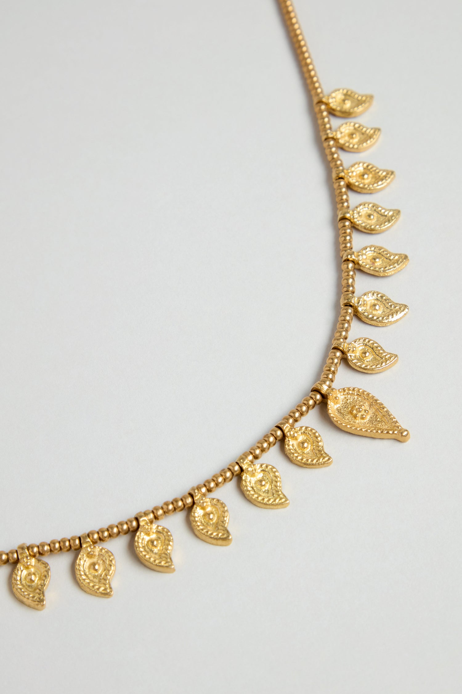 The Paisley Necklace features small paisley pendants in traditional Indian metalwork, evenly spaced on a beaded chain, and is shown against a light background.