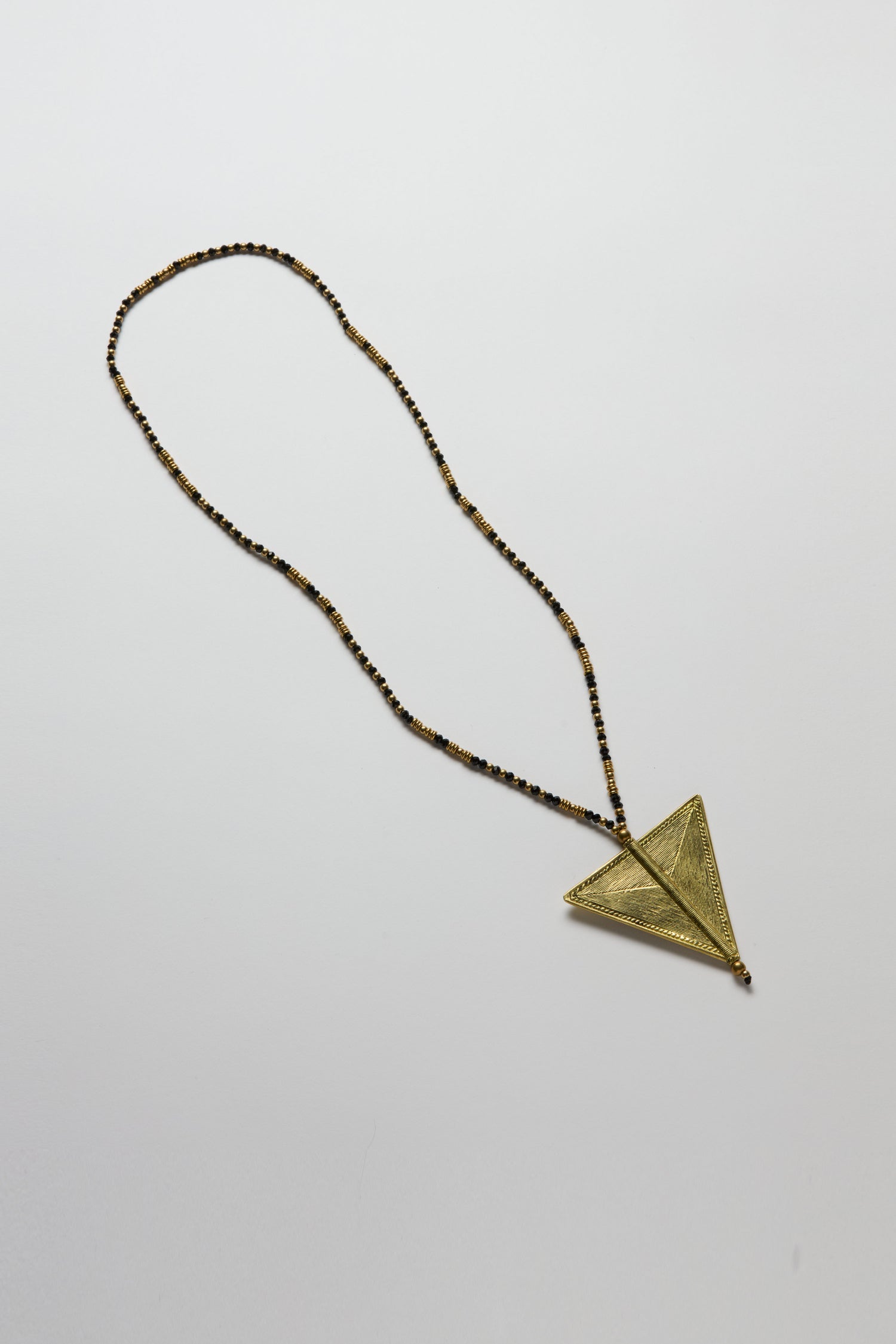 The Long Gold and Black Triangle Pendant from the Nataraj Collection is a beaded necklace featuring a textured, gold-colored, handcrafted triangle pendant, beautifully showcased on a plain white background.