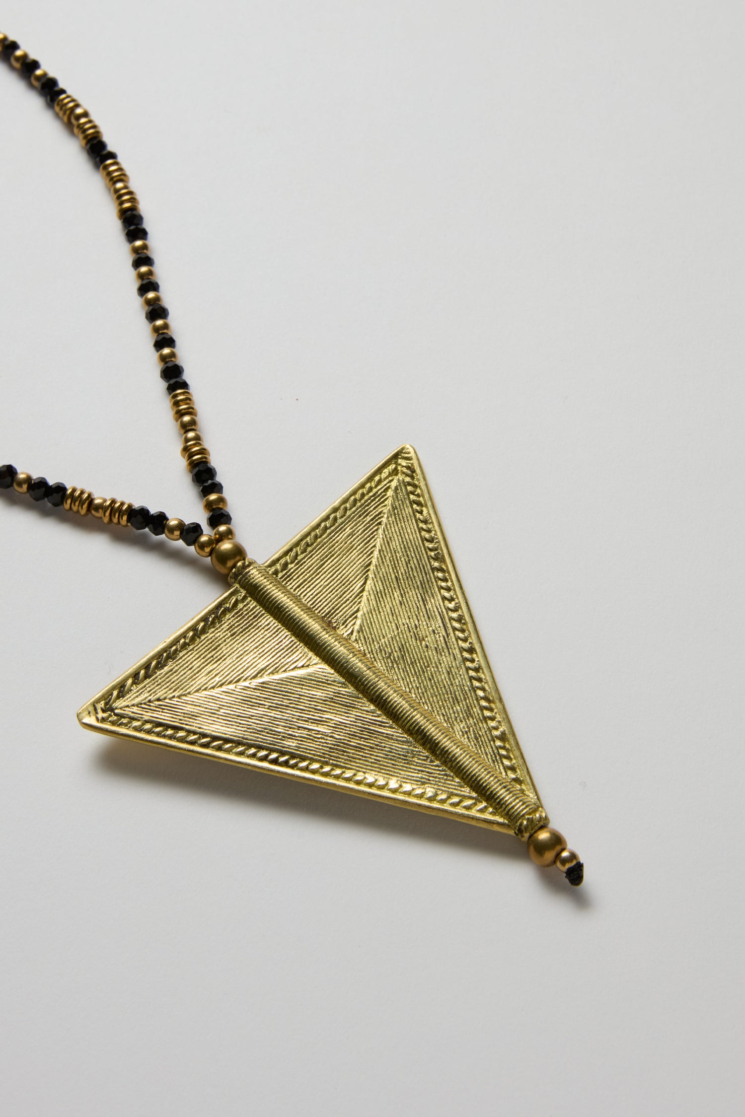 The Long Gold and Black Triangle Pendant from the Nataraj Collection features handcrafted geometric textured detailing and hangs on a beaded chain, displayed on a plain white background.