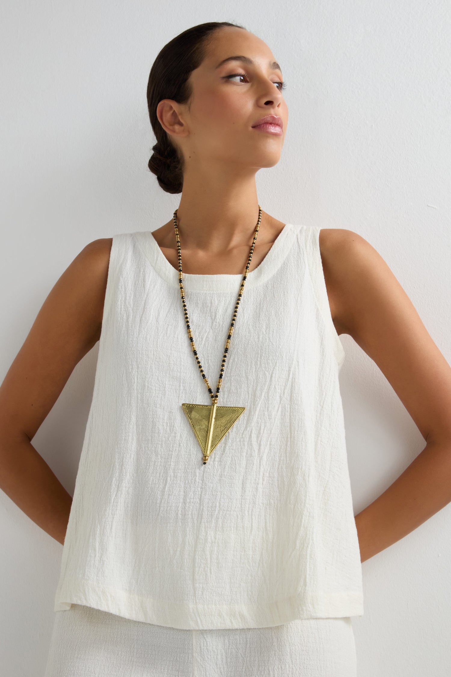 A person stands against a white wall wearing a sleeveless white top and pants, accessorized with the Long Gold and Black Triangle Pendant from the Nataraj Collection, inspired by tribal design.