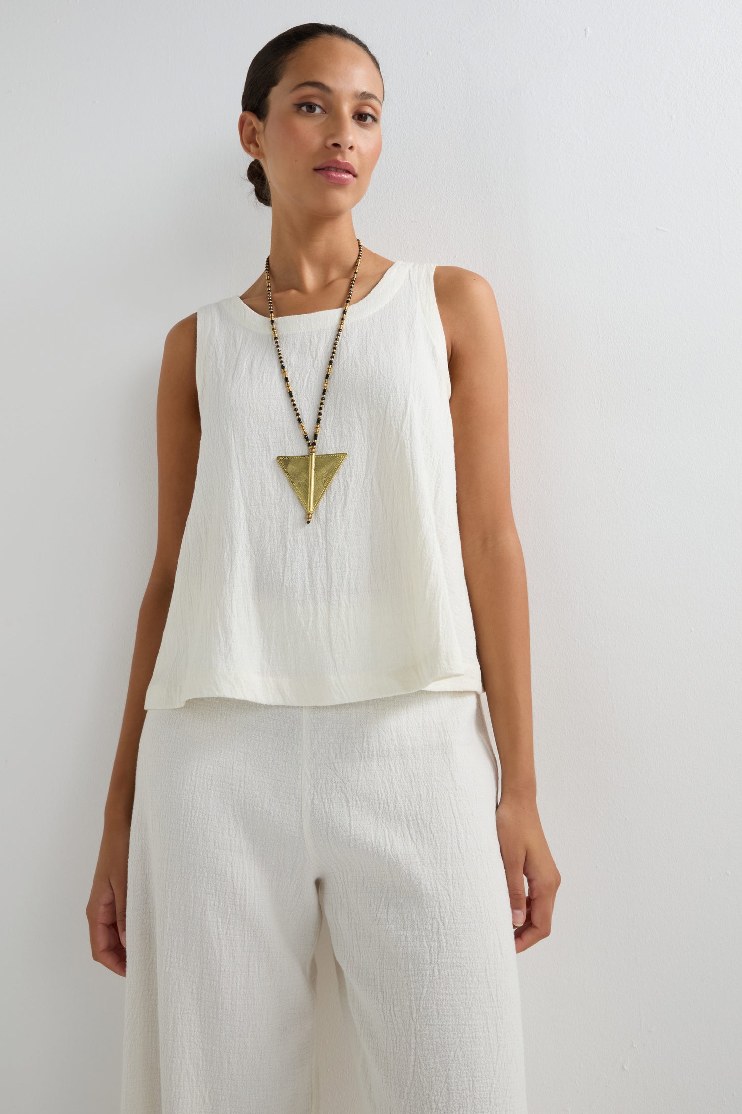 Woman in a sleeveless white top and pants, accessorized with the Long Gold and Black Triangle Pendant, featuring bold tribal design elements from the Nataraj Collection, standing against a plain white background.