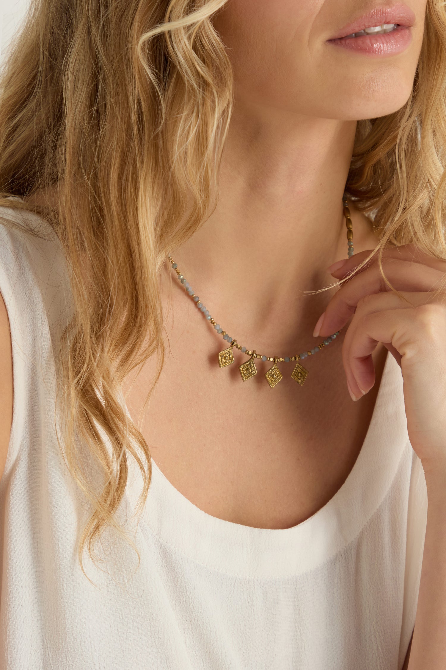 A woman with wavy blonde hair, in a sleeveless white top, wears the Eanna Necklace—an elegant piece featuring brass detailing and faceted cut glass beads—as she holds her hand near her chin.