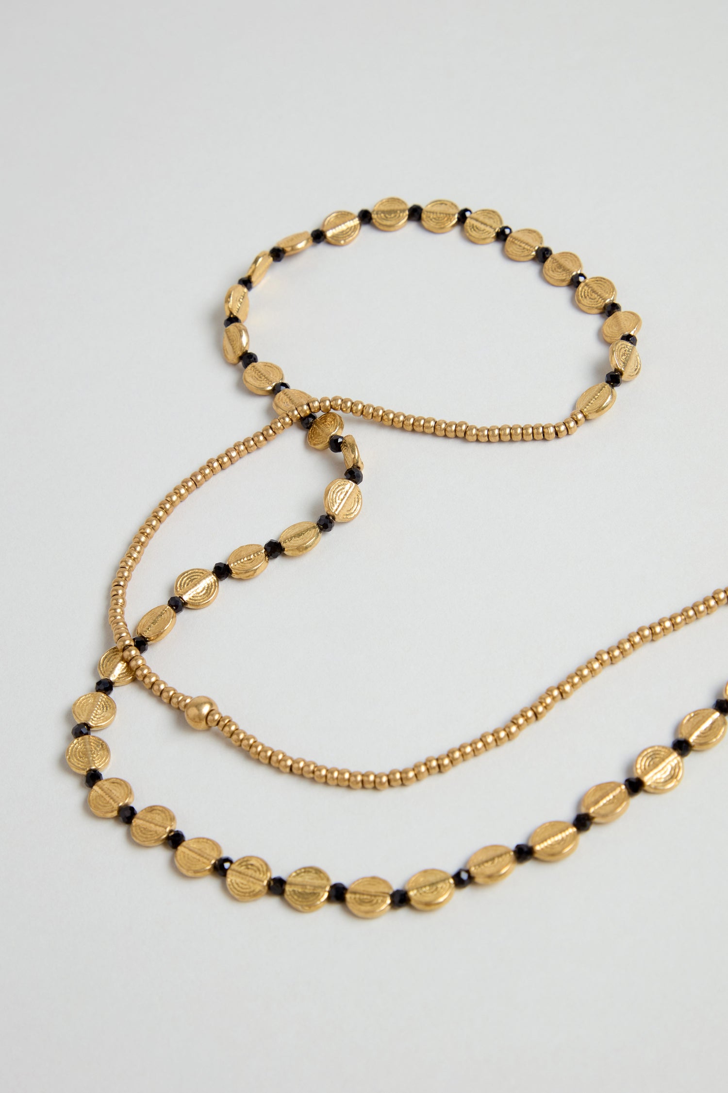 Two gold-toned beaded necklaces are displayed: one is a long brass bead strand, the other is the Mini Engraved Circles Bead Necklace featuring disc-shaped beads, black accents, and an engraved pendant.