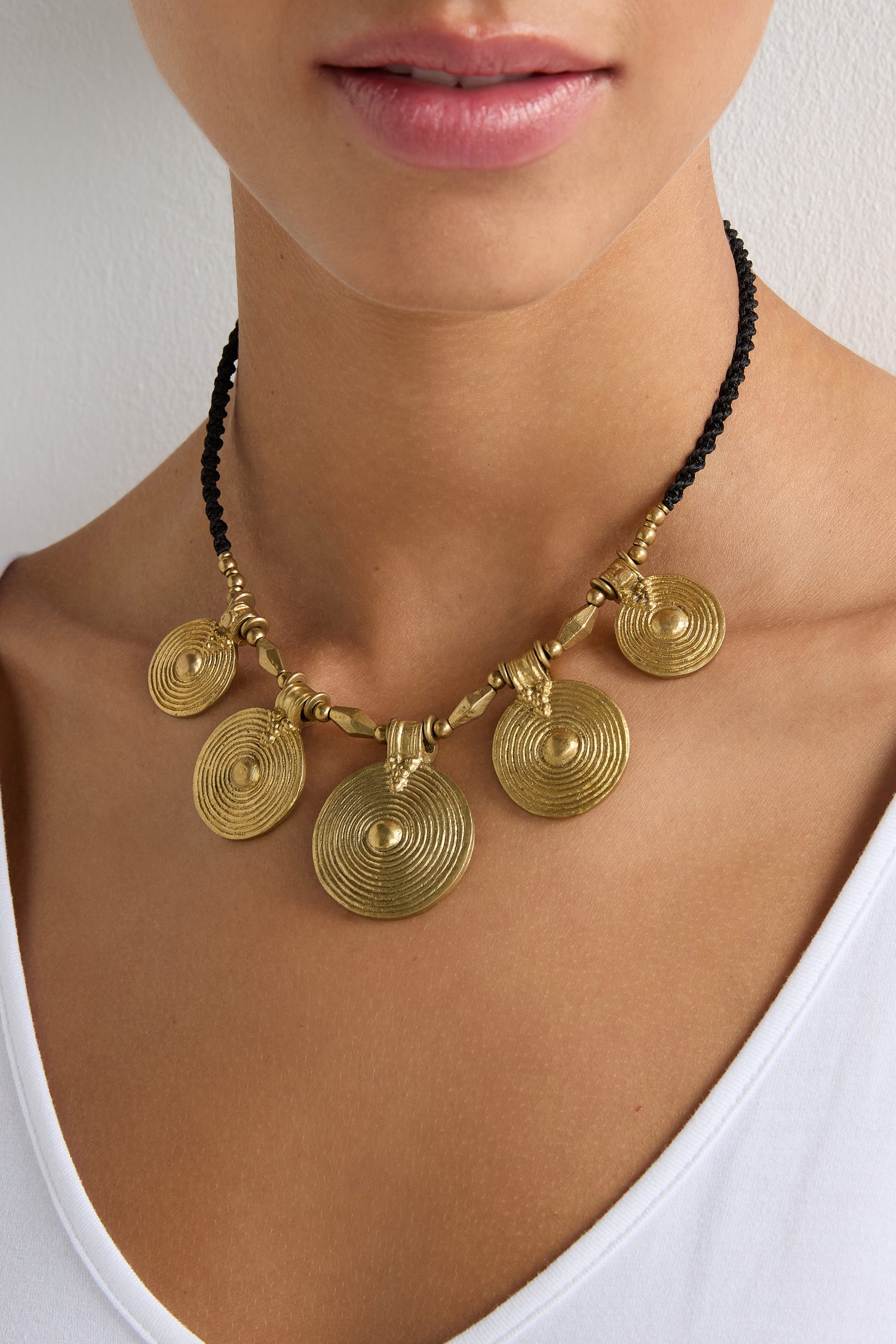 A person in a white top wears the Elissos Engraved Necklace, featuring five gold pendants on a black cord—highlighting contemporary elegance and exquisite handmade craftsmanship.