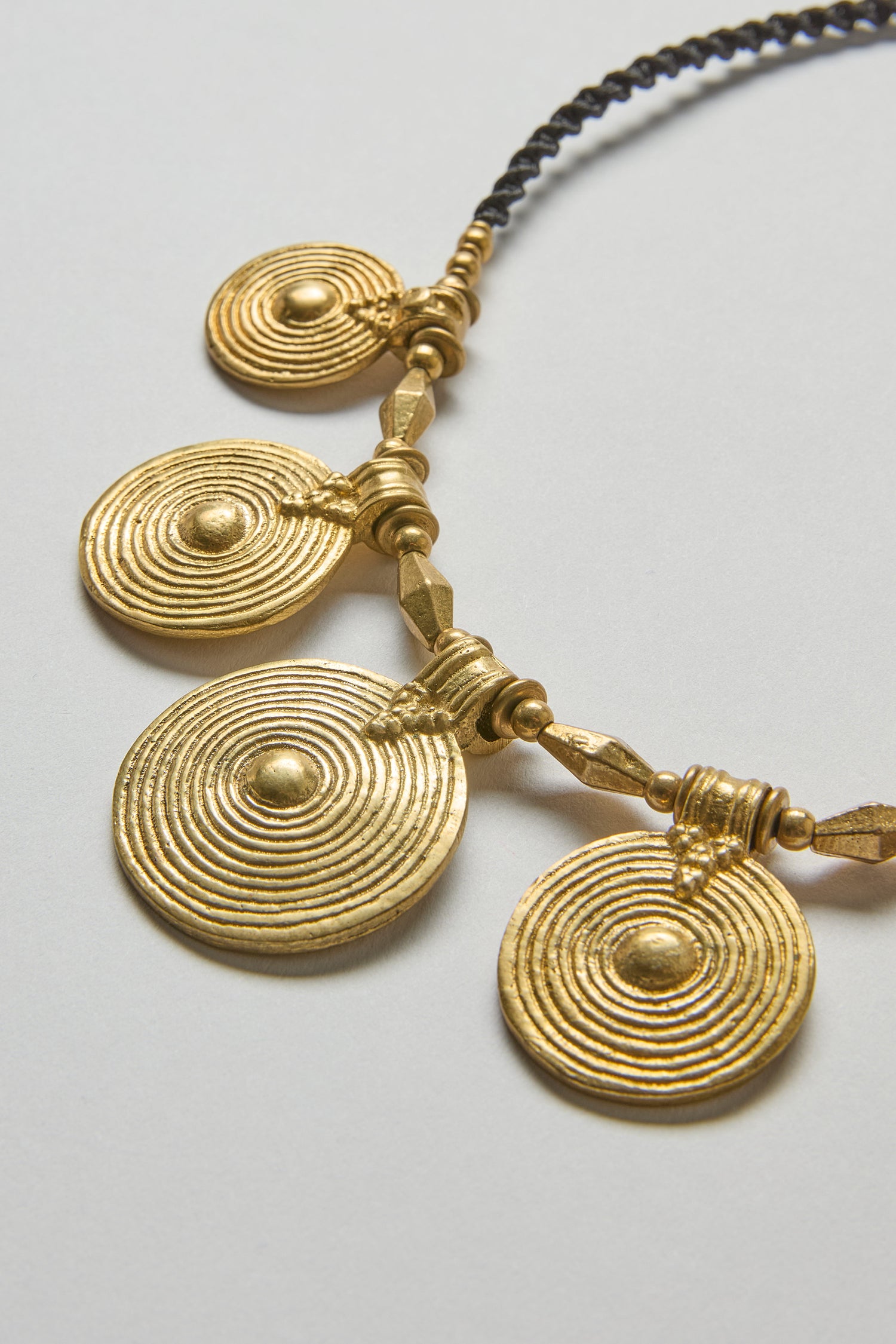 The Elissos Engraved Necklace features four large, round gold pendants with concentric circle designs on a black beaded string, combining handmade craftsmanship and modern style.